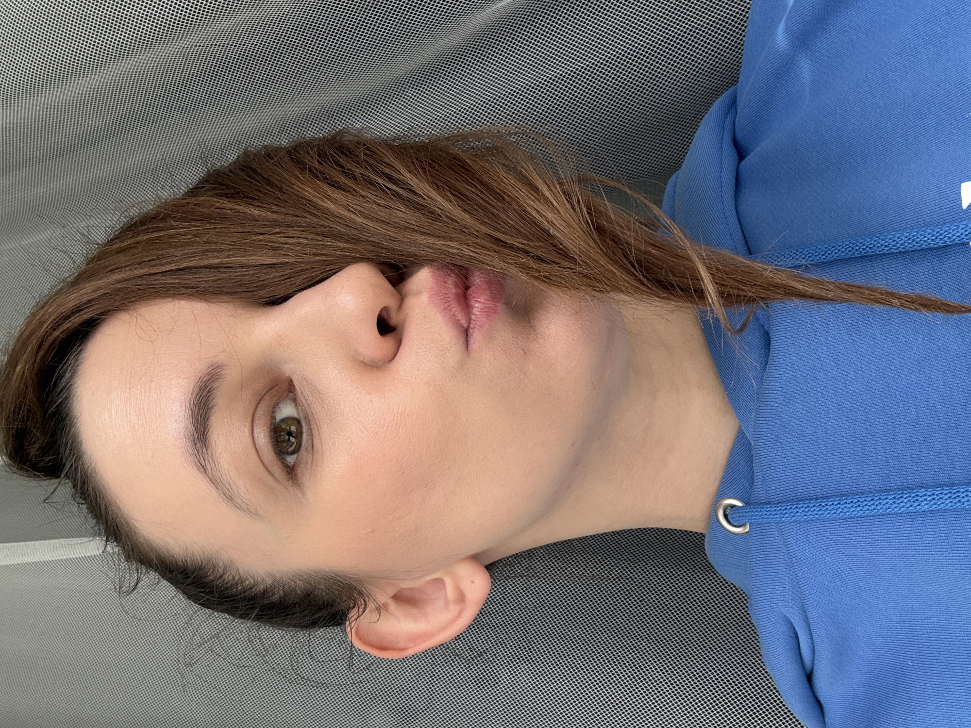 A close-up, tilted perspective image depicts a woman with light skin, brown hair, and light-colored eyes. She is wearing a blue hooded sweatshirt. The background appears to be a textured, possibly mesh, surface. The location is not identifiable as Chaikovsky, Russia, or any other specific location based on visual information.