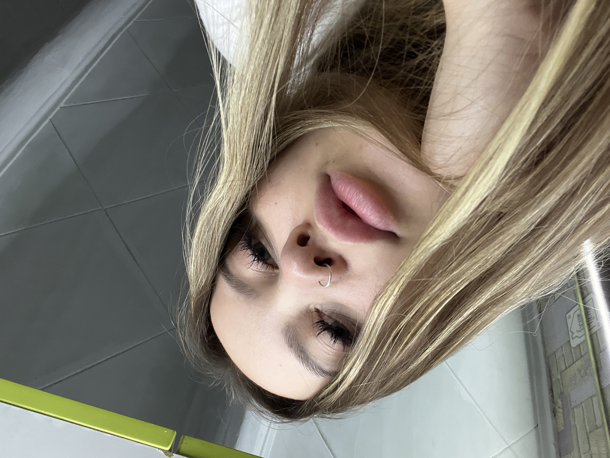 An upside-down portrait features a young woman with long, blonde hair, full lips, and dark, long eyelashes. A silver hoop nose ring is visible on her right nostril. Her eyes are cast downwards, suggesting she is looking into a camera positioned below her, capturing her from a low angle.
The background indicates an indoor setting, likely a room with tiled walls, possibly a bathroom. The left side of the image prominently displays dark grey square tiles with defined grout lines. On the right, lighter, possibly white or light grey, square tiles are visible. A bright green ledge or surface appears in the bottom left corner of the frame. Faintly visible on the right, behind her hair, is a reflection or a decorative item displaying the word "Home." The woman's white clothing, possibly a top, is visible near her head. The scene is located in Port-Katon, Russia.