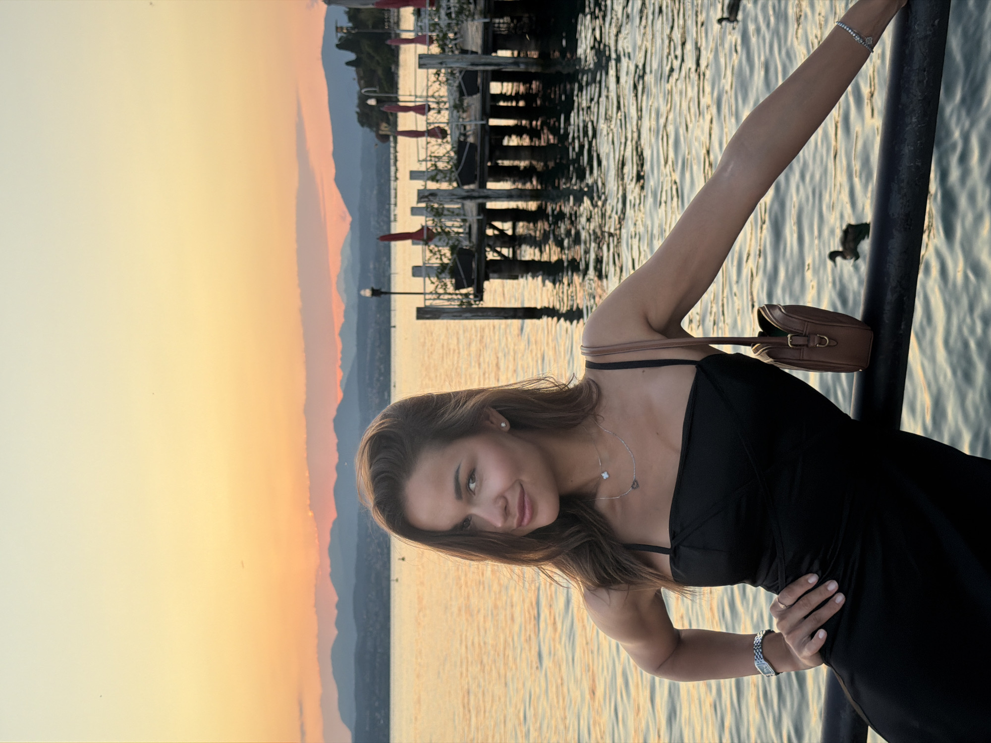A young woman with long brown hair and a black dress is posing on a railing in front of a body of water at sunset. The water is calm and reflects the orange and yellow hues of the sky. In the background, there is a dock with red umbrellas and a distant shoreline with mountains. The woman is looking at the camera with a slight smile, and she has a brown purse slung over her shoulder. She is wearing a silver necklace and a watch. The scene is serene and picturesque, evoking a sense of relaxation and enjoyment. The location is believed to be Sirmione, Italy, a popular tourist destination known for its beautiful lake views and historic charm.