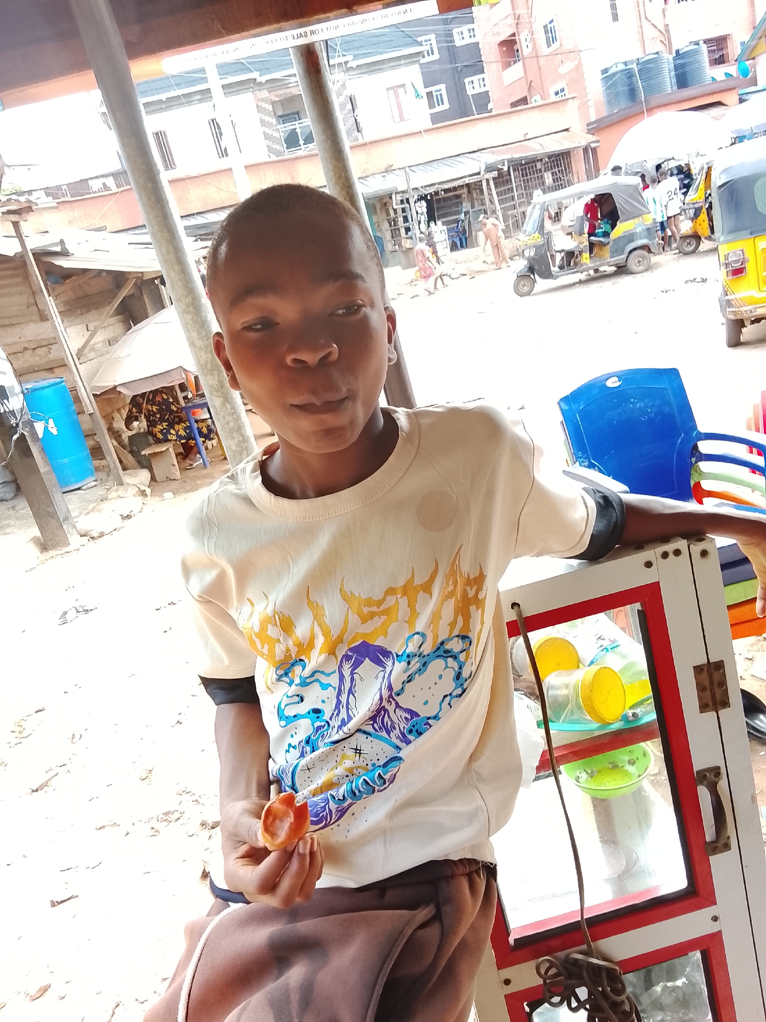 A young boy, likely of African descent, is featured prominently in the foreground of a bustling outdoor street or market scene. He wears a light-colored t-shirt adorned with a vibrant graphic design in yellow, purple, and blue hues, and dark shorts or pants. With short hair and a neutral expression, he looks slightly away from the camera while holding a reddish-orange fruit in his right hand, appearing to be in the midst of eating it. An earring is visible in his left ear.
To the boy's right, a white and red display case or stall is present, with yellow-lidded containers and a green bowl visible inside. A black cord hangs from the side of this unit. On his left, a bright blue plastic barrel is partially seen.
The background unfolds into a lively street view under what appears to be sunny daytime conditions. The ground is unpaved and dusty. Multiple auto-rickshaws (tricycles, commonly known as Keke Napep), some yellow and others black/yellow, are parked or moving on the road. Various multi-story buildings, some with exposed concrete and others painted, line the street, with water tanks visible on several rooftops. Numerous market stalls or small shops with metal grates or open fronts are also part of the landscape. Other people are visible in the background, engaged in daily activities, including one person wearing a white jersey with the number "24". Faint, inverted text "HOT FOR SALE NOT FOR" and "10" is discernible on a distant sign, possibly a reflection. The setting suggests a vibrant, everyday urban or semi-urban environment, characteristic of a warm climate.