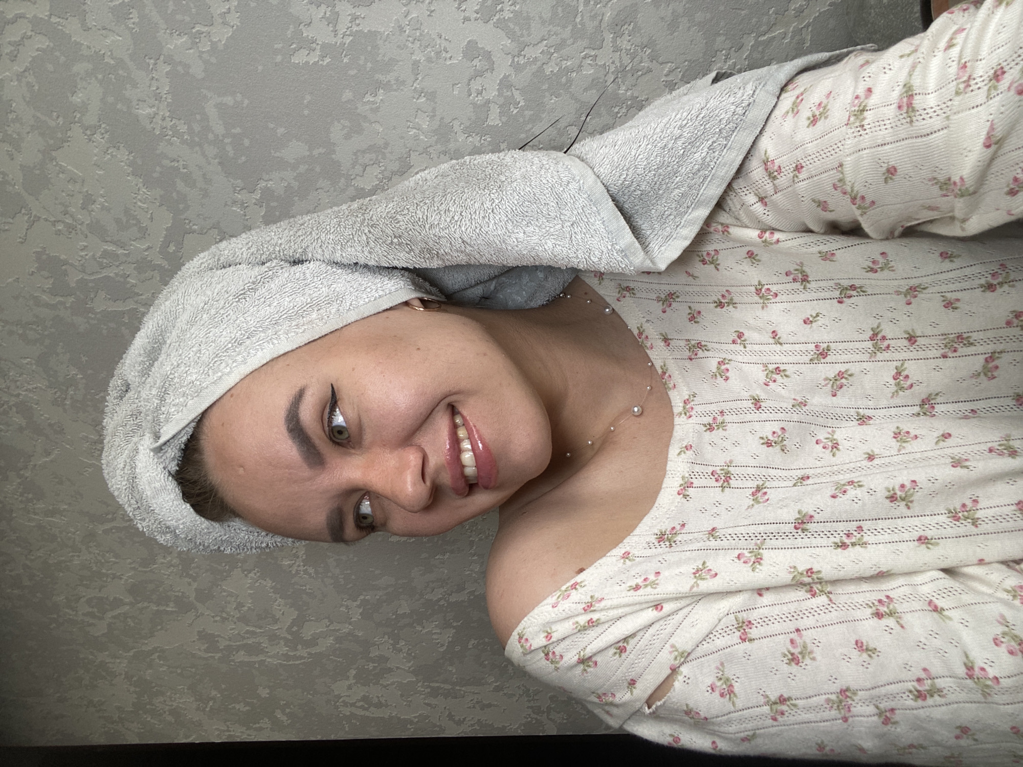 A young woman with a towel wrapped around her head is looking to her left and smiling. She is wearing a light-colored, floral-patterned shirt with an off-the-shoulder design and a delicate pearl necklace. Her makeup includes winged eyeliner and lip gloss. The background is a textured gray wall. The image was taken indoors, in a residential setting, likely in Chaikovsky, Russia, as indicated by the context.