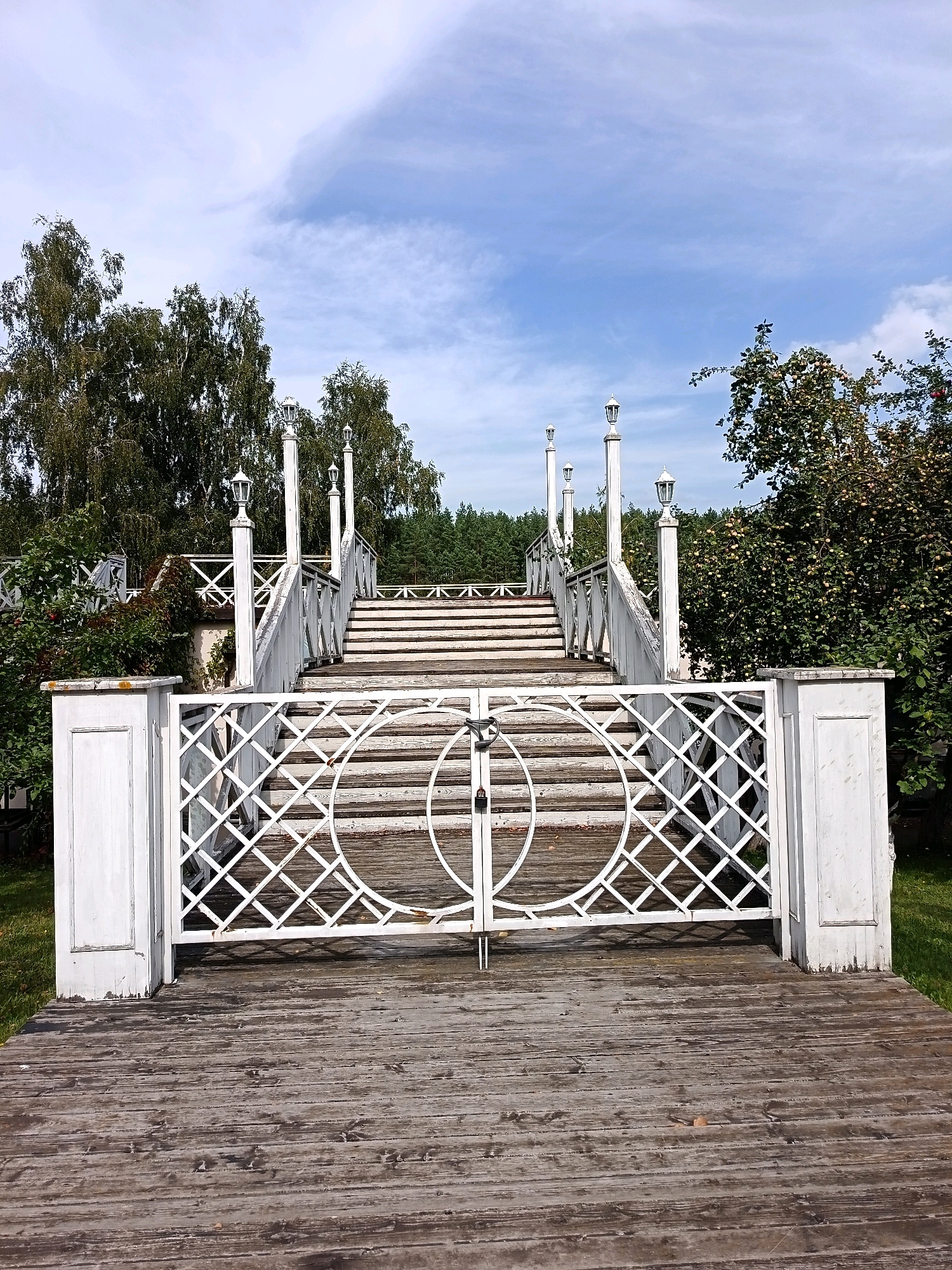 The image depicts an outdoor scene featuring a white wooden staircase with ornate railings and decorative lamps, leading upwards. At the base of the staircase, there is a closed white gate with a lattice design and circular accents, secured by a padlock. The stairs and a wooden deck are in the foreground. Lush greenery, including trees and bushes, surrounds the structure, with a forest visible in the background. The sky above is partly cloudy. The setting appears to be a park or garden area, possibly within the city of Kondyreva, Russia. There are no people or animals present in the image.
