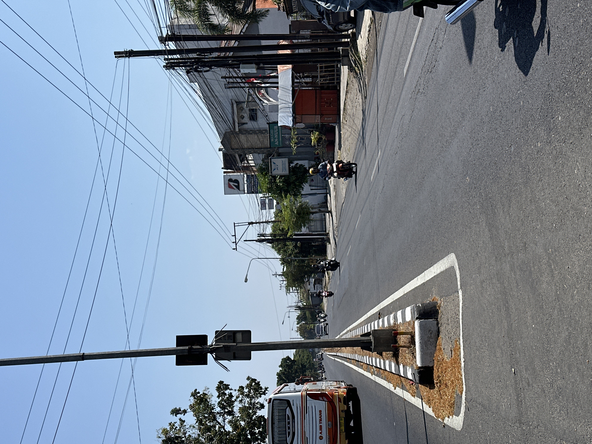 The image depicts a street scene in Bulakrejo, Indonesia, on a clear, sunny day. A white bus with orange and red accents, identified as "AJIB TRANS" and "PARIWISATA," travels in the left lane. A traffic pole with multiple signs is positioned in the median. Several motorcycles are also present on the road, with one rider wearing a yellow helmet and another wearing a blue helmet and jacket. On the right side of the road, buildings line the street, with one featuring a "Bridgestone" sign and another with "MASJID AN-NUUR" signage. Power lines crisscross the sky above. The road surface appears to be asphalt, with white lane markings and a striped curb separating the lanes and median. Debris, possibly dried leaves or seeds, is scattered on the median.