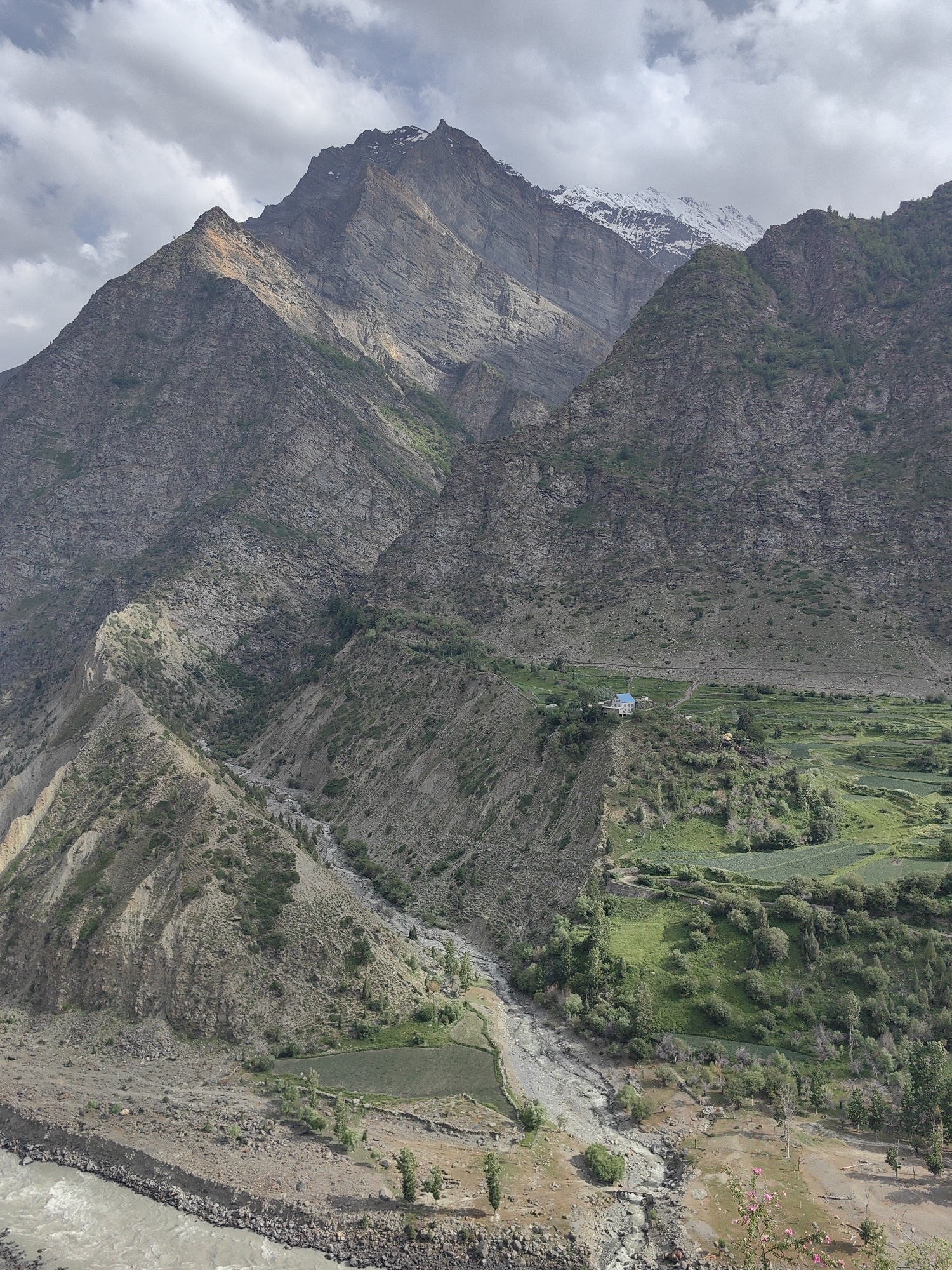 The image shows a mountainous landscape with terraced fields and a river. In the middle ground, a cluster of buildings with blue roofs is visible on a hillside, possibly indicating a village or settlement. The mountains are rocky and steep, with some vegetation on their slopes. The sky is partly cloudy. This scene is consistent with the scenery found in Kirting, India, known for its mountainous terrain and agricultural practices.