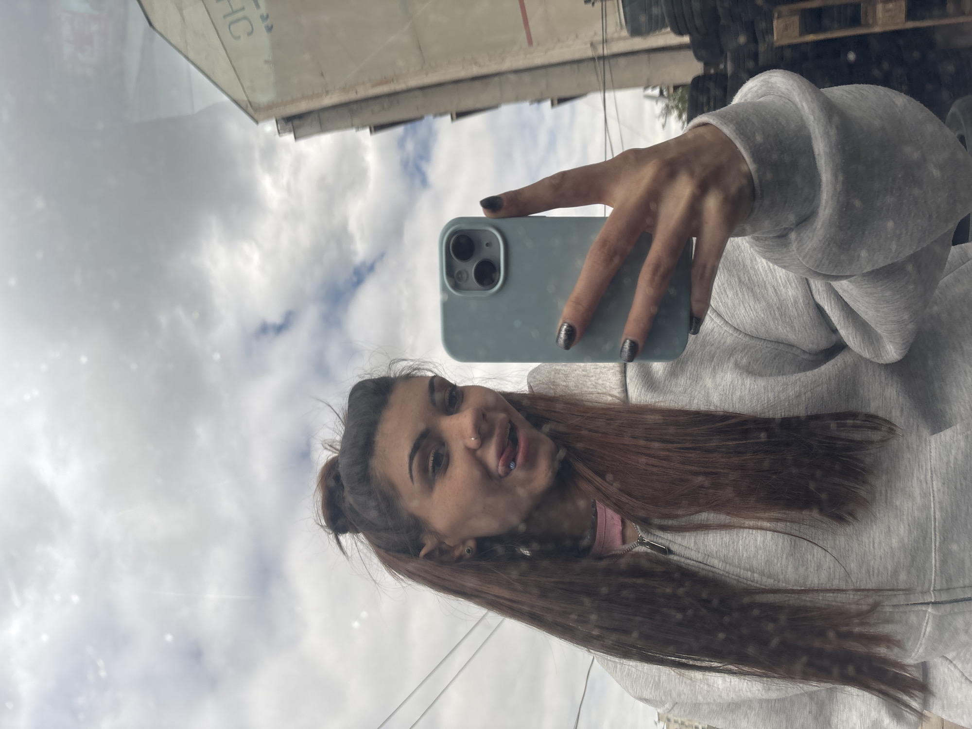 A young woman is taking a selfie using a smartphone. She's reflected in a window, which also reflects a building exterior and cloudy sky. The woman has brown hair, partly tied up, with a nose piercing and a blue tongue piercing. She's wearing a gray hoodie. The phone she is holding is light blue. The background reflects a building and a stack of tires, potentially indicating an industrial area. Without additional information, I am unable to confirm this is in Sofia.