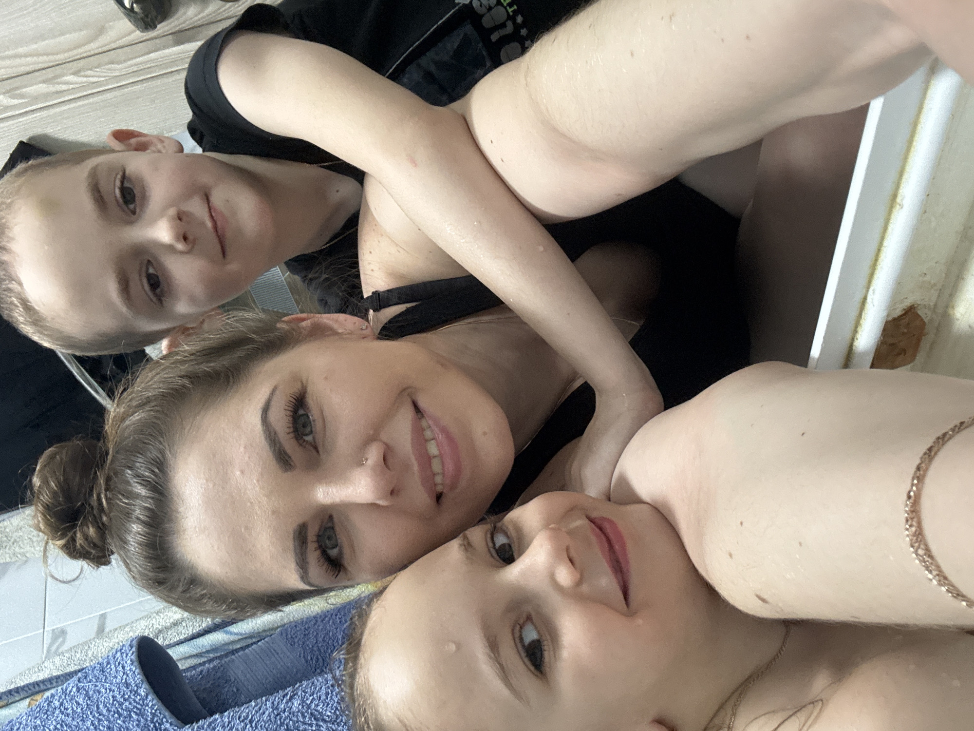 The image appears to be a selfie taken by a woman with two children, likely in a bathroom. The woman is in the center of the frame, smiling. A young boy is positioned above her, and a younger girl is below. All three subjects appear damp, possibly indicating they have just taken a bath or shower. The bathroom setting is suggested by the presence of blue towels, tiled walls, and a bathtub. The location mentioned, Раздолье, Russia, cannot be verified from the image's content.