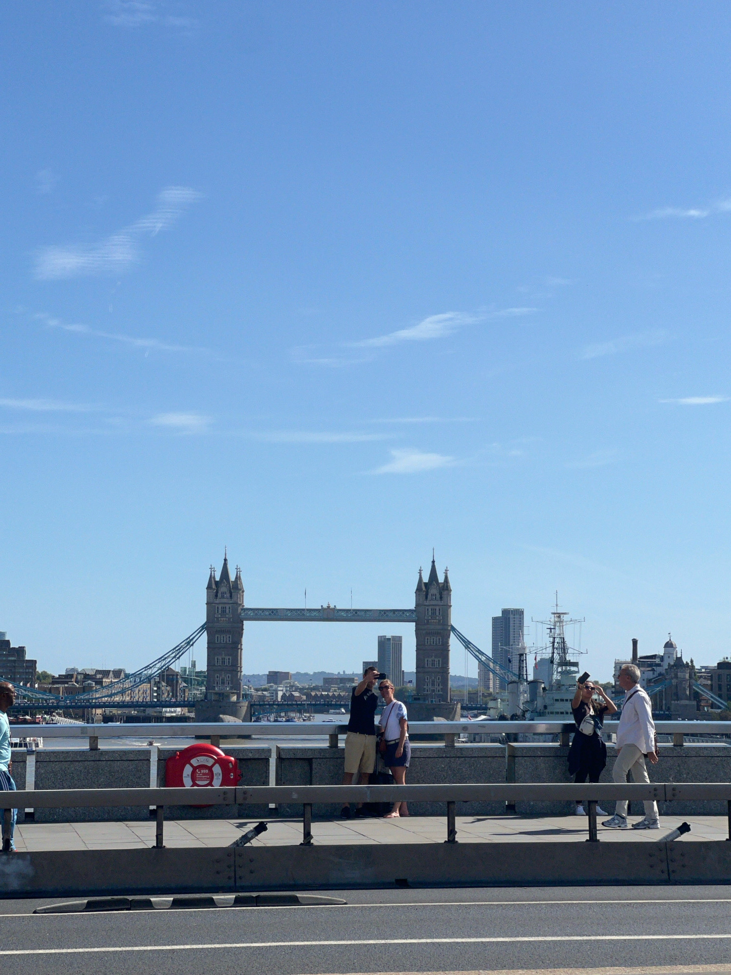 This image shows a view of Tower Bridge in London, United Kingdom, under a clear blue sky with scattered wispy clouds. In the foreground, a man in a light blue shirt and dark pants stands on a bridge sidewalk. To his right, a red lifebuoy with "999 Full Emergency" printed on it is mounted on a wall. Further to the right, a couple stands close together, with the woman taking a photo of the man with her phone. Behind them, another couple walks away from the camera, the woman in a white shirt and dark skirt, and the man in a white shirt and light-colored pants. In the background, Tower Bridge is prominently visible with its iconic towers and suspension structure. The River Thames flows beneath the bridge, with boats and the London skyline visible in the distance, including modern skyscrapers and other historic buildings. A large ship, possibly a museum vessel, is docked near the bridge. The overall scene depicts a sunny day with tourists enjoying the iconic London landmark.
