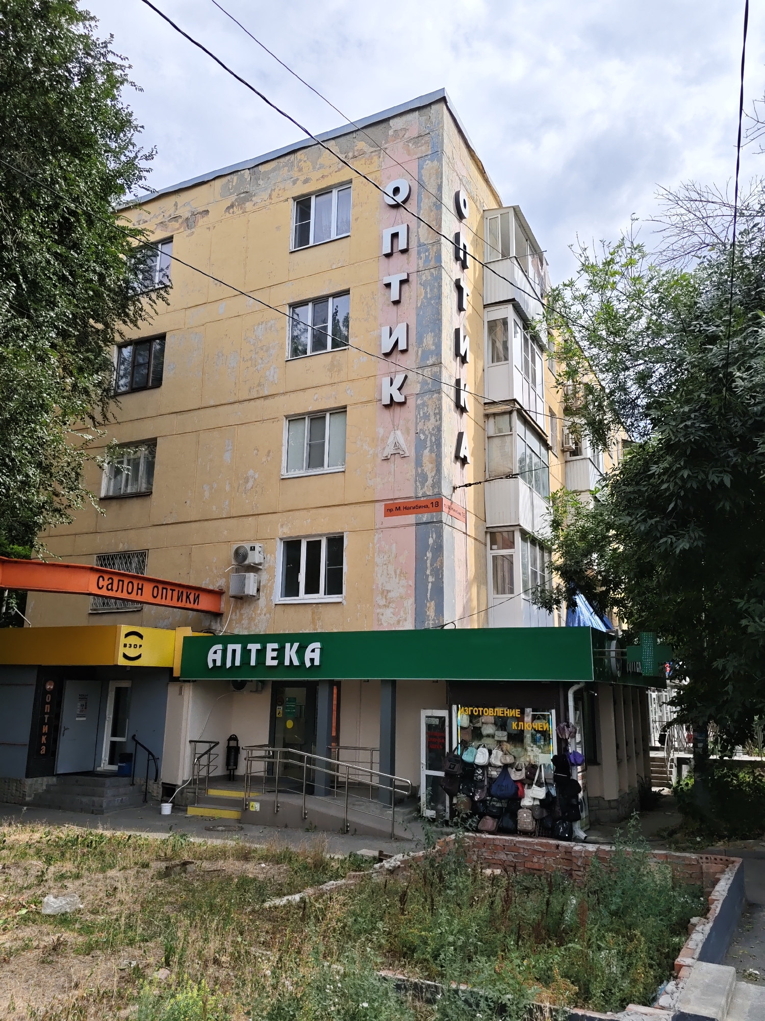 The image shows a multi-story apartment building with commercial establishments on the ground floor in Rostov-on-Don, Russia. The building has a faded yellow facade with peeling paint, and some sections are painted with vertical stripes of pink and blue. There are several windows and balconies on the upper floors.
On the ground floor, there are two visible storefronts. The left one is for "САЛОН ОПТИКИ" (Optics Salon) with an orange sign. To its right is a storefront labeled "АПТЕКА" (Pharmacy) with a green awning. Next to the pharmacy, there is a small shop displaying numerous handbags and a sign that says "ИЗГОТОВЛЕНИЕ КЛЮЧЕЙ" (Key Making).
The area in front of the building appears to be a sidewalk with some stairs and a ramp for accessibility. There is vegetation growing in some areas, including trees lining the building and overgrown weeds in a partially constructed brick enclosure in the foreground. Overhead, power lines are visible against a cloudy sky. The general impression is of an older urban environment.