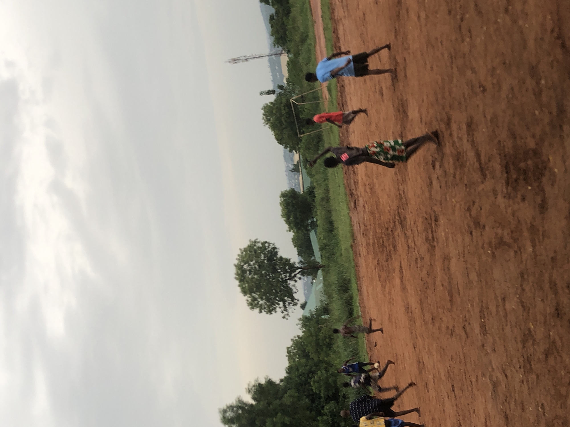 The image displays an outdoor scene in Ukwu-Abwa, Nigeria, featuring several young males on a reddish-brown dirt field, characteristic of laterite soil. The primary activity appears to be a game of football (soccer).
In the mid-ground, a boy in a blue shirt and black shorts stands near a rudimentary, white-framed goalpost. Another boy in a red shirt is positioned closer to the goal. To their right, a boy wearing a patterned (possibly floral or camouflage) shirt and shorts is captured in a dynamic pose, suggesting movement during play. Further down the field, other figures are visible, participating in the group activity.
The setting includes a backdrop of lush green trees and foliage, indicating a natural or semi-natural environment. A communication tower or mast is discernible in the distant background, hinting at a blend of rural and modern infrastructure. The sky is heavily overcast, suggesting either a cloudy day or the late afternoon/early evening hours, as the light is diffused and lacks harsh shadows.
No visible text is present in the image. The overall impression is one of children or young adults engaged in recreational sports in a community setting.