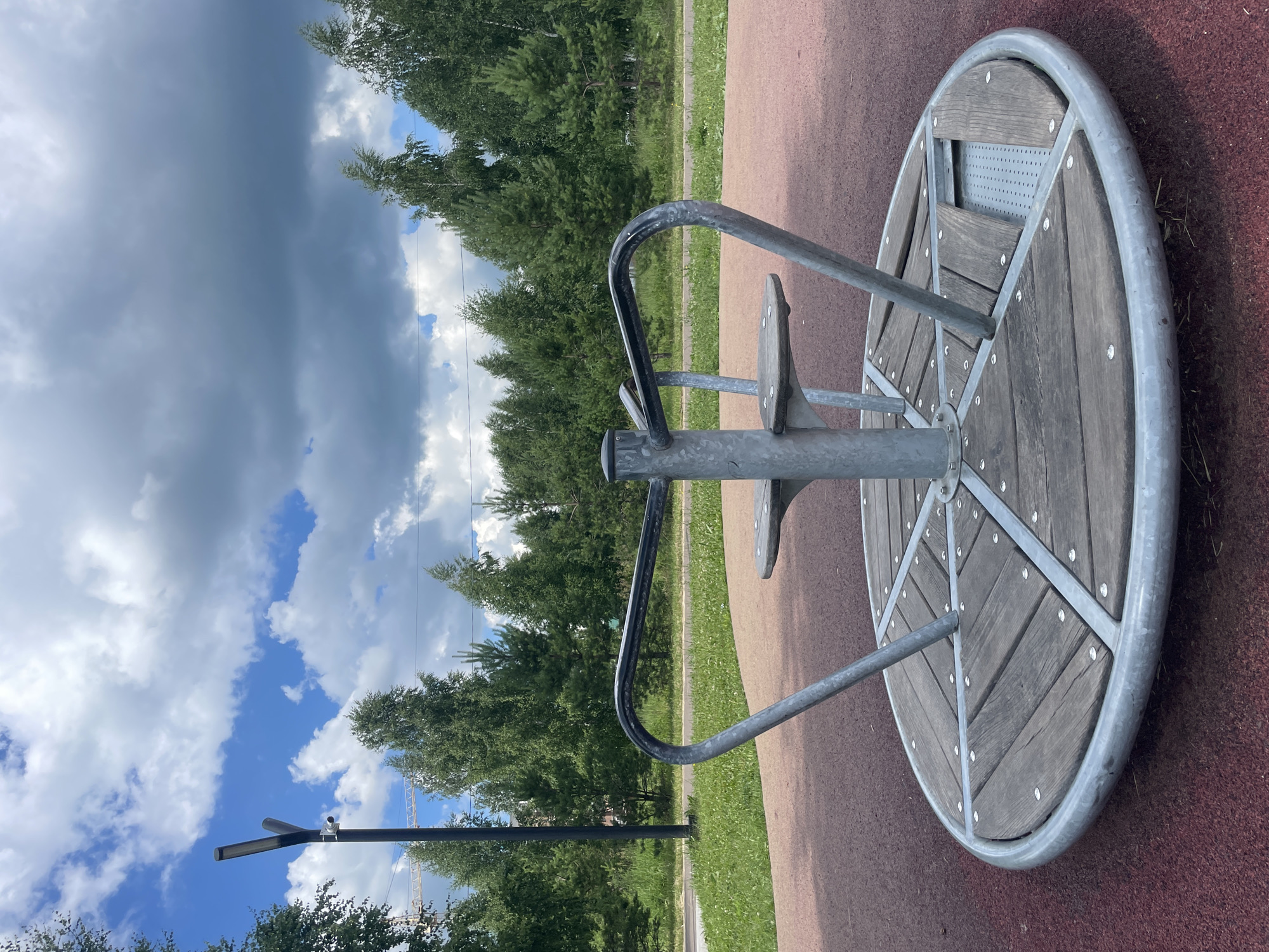 A playground merry-go-round, constructed from grey metal and weathered wood planks, stands on a rubberized, maroon-colored surface. The merry-go-round features a central metal pole with two attached seats or platforms. The scene is set against a backdrop of green trees and a sky with dramatic, cloudy formations. A modern lamppost with a security camera is visible to the left, and construction cranes are present in the distance, suggesting ongoing development. The overall environment appears to be a park or recreational area in Laishevo, Russia.