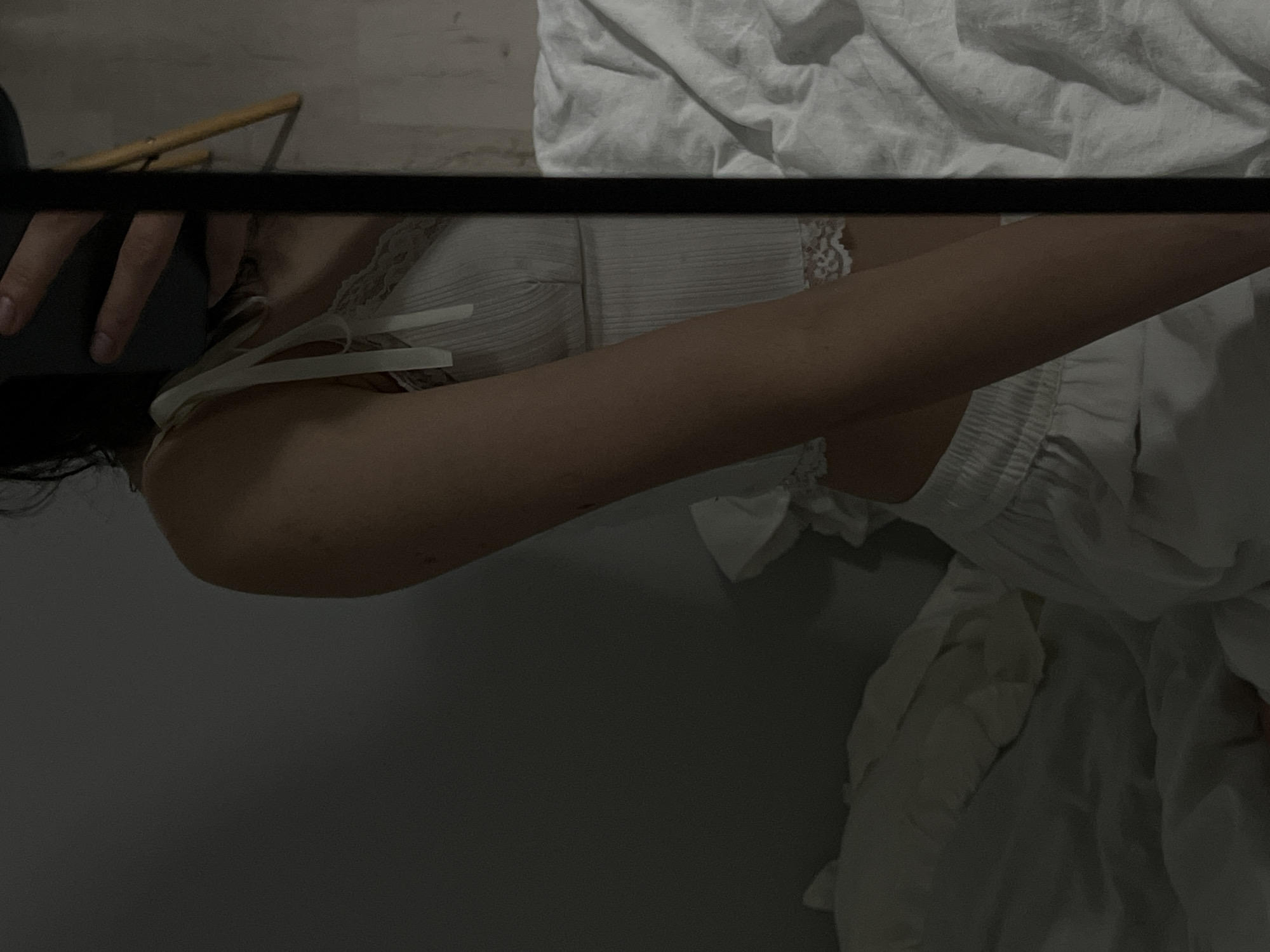 The image shows a person, likely a young woman, sitting on a bed in a dimly lit room. The person is wearing white pajamas, consisting of a ribbed tank top with lace trim and wide-legged pants. Her arm and shoulder are visible, and she appears to be holding a phone in her hand, possibly taking a mirror selfie. The bed is covered with white linen, and the room has a simple, minimalist aesthetic. The background shows a plain wall and a hint of a wooden chair. The overall impression is one of a private, intimate moment. The location is not specified within the image or its metadata, and therefore cannot be determined to be Ternopil, Ukraine.