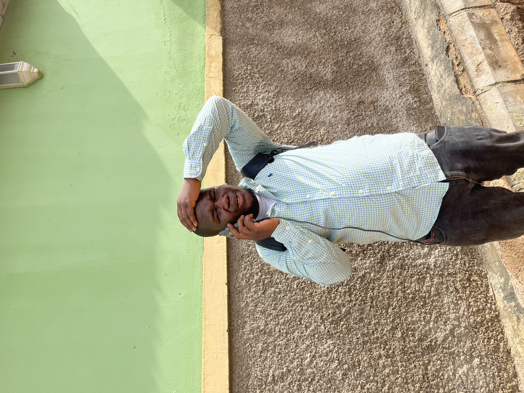 A Black man stands against a textured, brown wall under a bright, lime-green painted section. He is wearing a light blue and white checkered shirt, dark grey pants, and a black backpack. The man is holding a dark-colored smartphone to his right ear with his right hand and has his left hand on the back of his head, with his eyes closed, appearing distressed. A yellow band separates the green and brown sections of the wall. A white outdoor light fixture is mounted on the upper right portion of the green wall. The scene appears to be outdoors, possibly on a street in Jos, Nigeria, given the architectural style and colors.