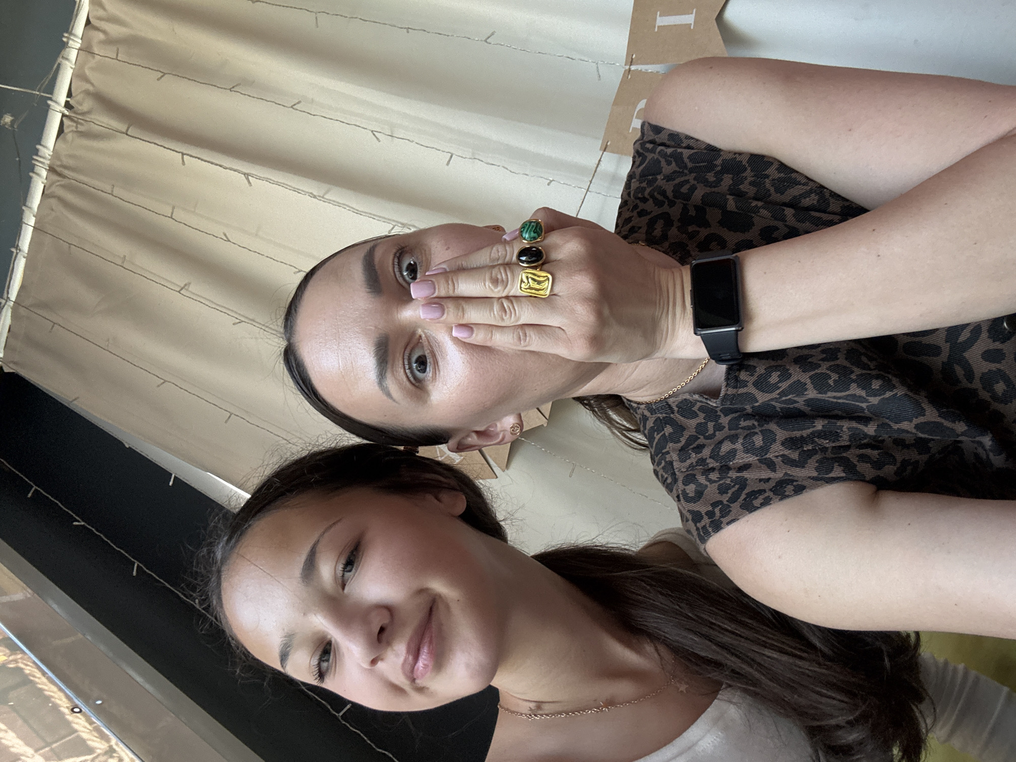 The image shows a medium shot of two women posing for a selfie. The woman on the left has long, dark hair and is smiling. The woman on the right is covering her mouth with her hand, revealing several rings on her fingers. The background appears to be an indoor setting with draped fabric and string lights. There is no discernible information in the image to suggest a location in Sochi, Russia.