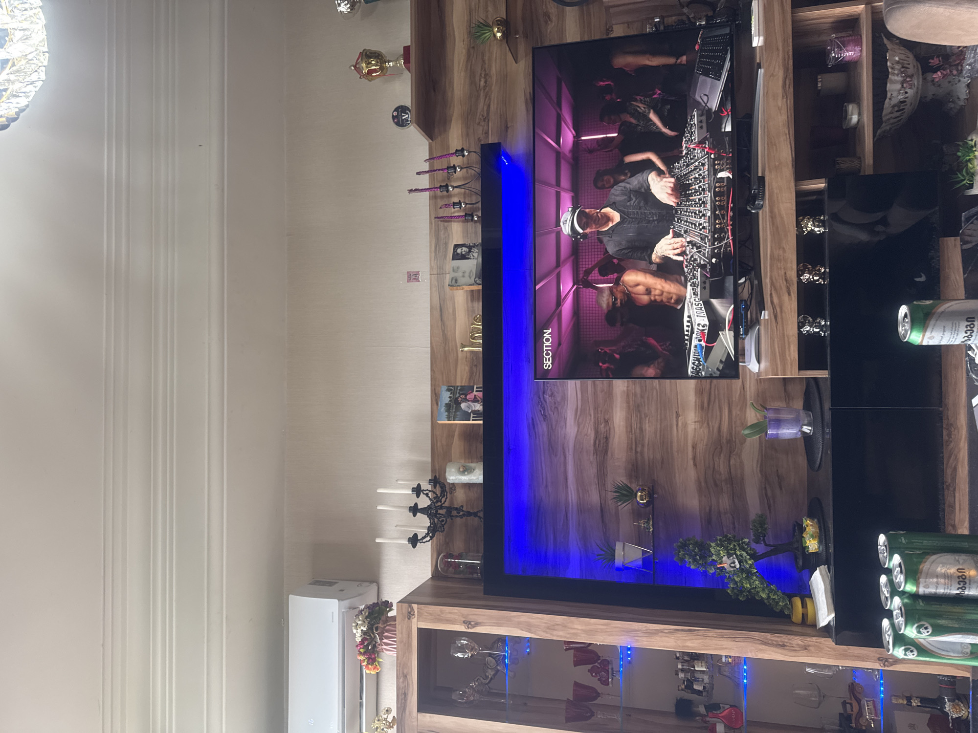 The image shows a living room with a large television displaying a DJ performing at a club. The room features wooden furniture, including a display cabinet filled with glassware and decorative items. There is an air conditioning unit mounted on the wall above the cabinet. On a shelf above the television, there are several decorative objects, including candles and trophies. In the foreground, there are several cans of beer. The overall impression is that of a modern, well-decorated living space. The details in the image, such as the design of the furniture and the electronics, suggest a contemporary urban setting. While the image does not explicitly state the location, the presence of Georgian script on the beer cans strongly indicates that the scene is in Tbilisi, Georgia.