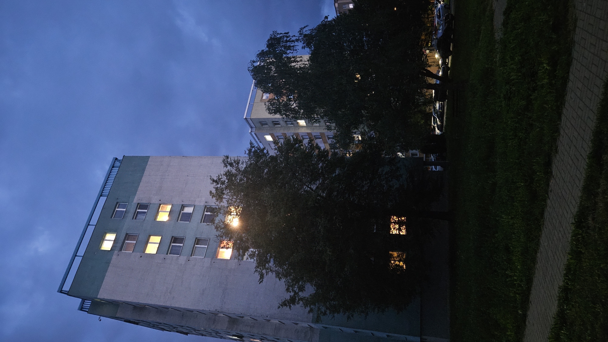 The image depicts a nighttime scene in the city of Prešov, Slovakia. The foreground features a paved walkway bordering a dark grassy area. In the midground, two trees stand prominently, their foliage obscuring parts of the background. Beyond the trees, two apartment buildings are visible. The building on the left is taller and appears to be a high-rise, with several windows illuminated from within. The building on the right is shorter, also showing lit windows. The sky above is dark and cloudy, suggesting either dusk or night. Parked cars are faintly visible in the distance behind the trees and in front of the shorter apartment building. The overall atmosphere is quiet and urban.