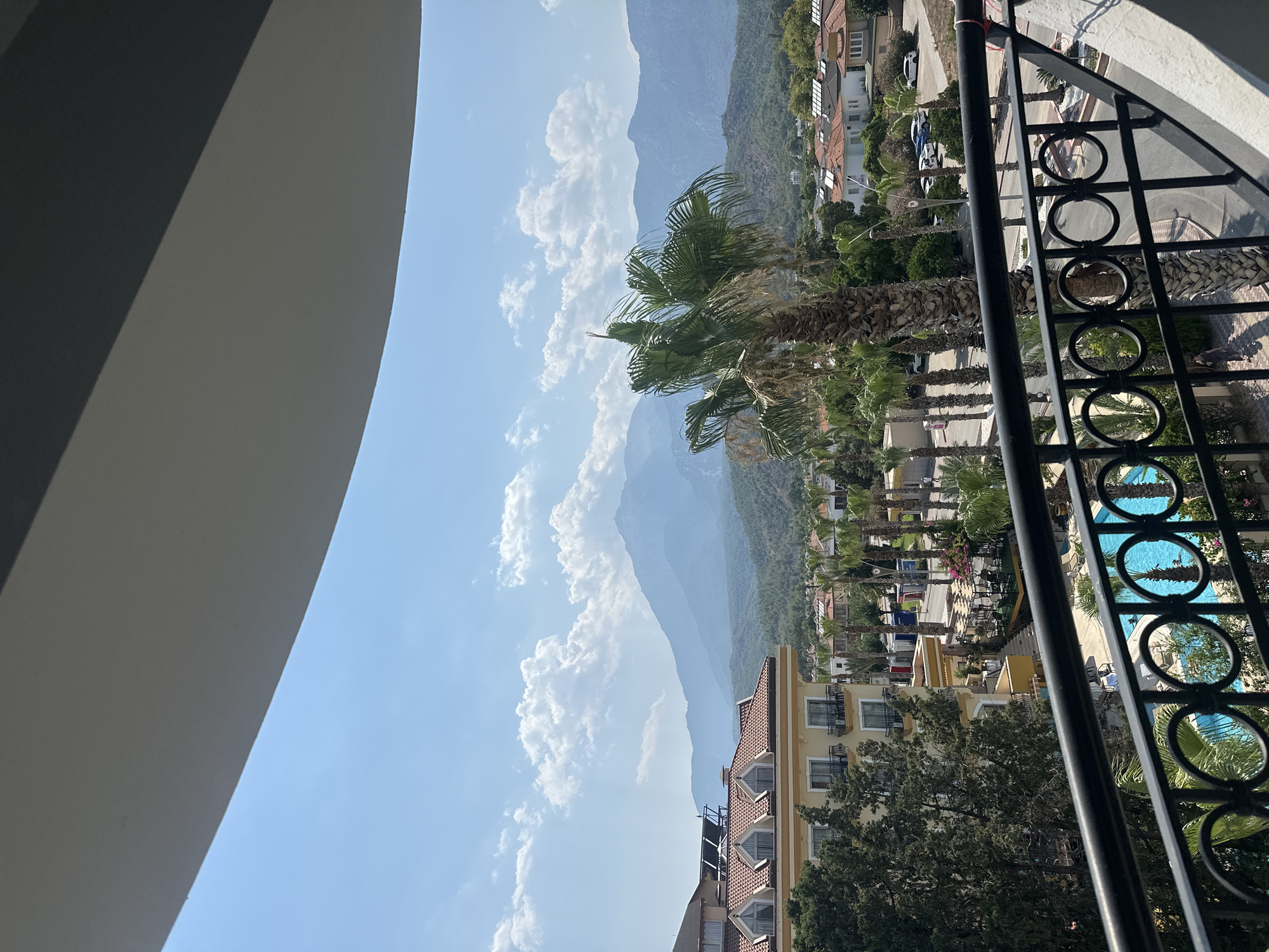 This image captures a scenic view of Kemer, Turkey, from a balcony. In the foreground, a black metal railing with circular embellishments is visible, partially obscuring a bright blue swimming pool below. Beyond the pool, palm trees line a street with parked cars and several buildings. One prominent building is a yellow hotel with a red tiled roof and multiple balconies. In the background, lush green hills rise to meet a hazy mountain range under a clear blue sky dotted with white clouds. The overall impression is one of a sunny resort town.