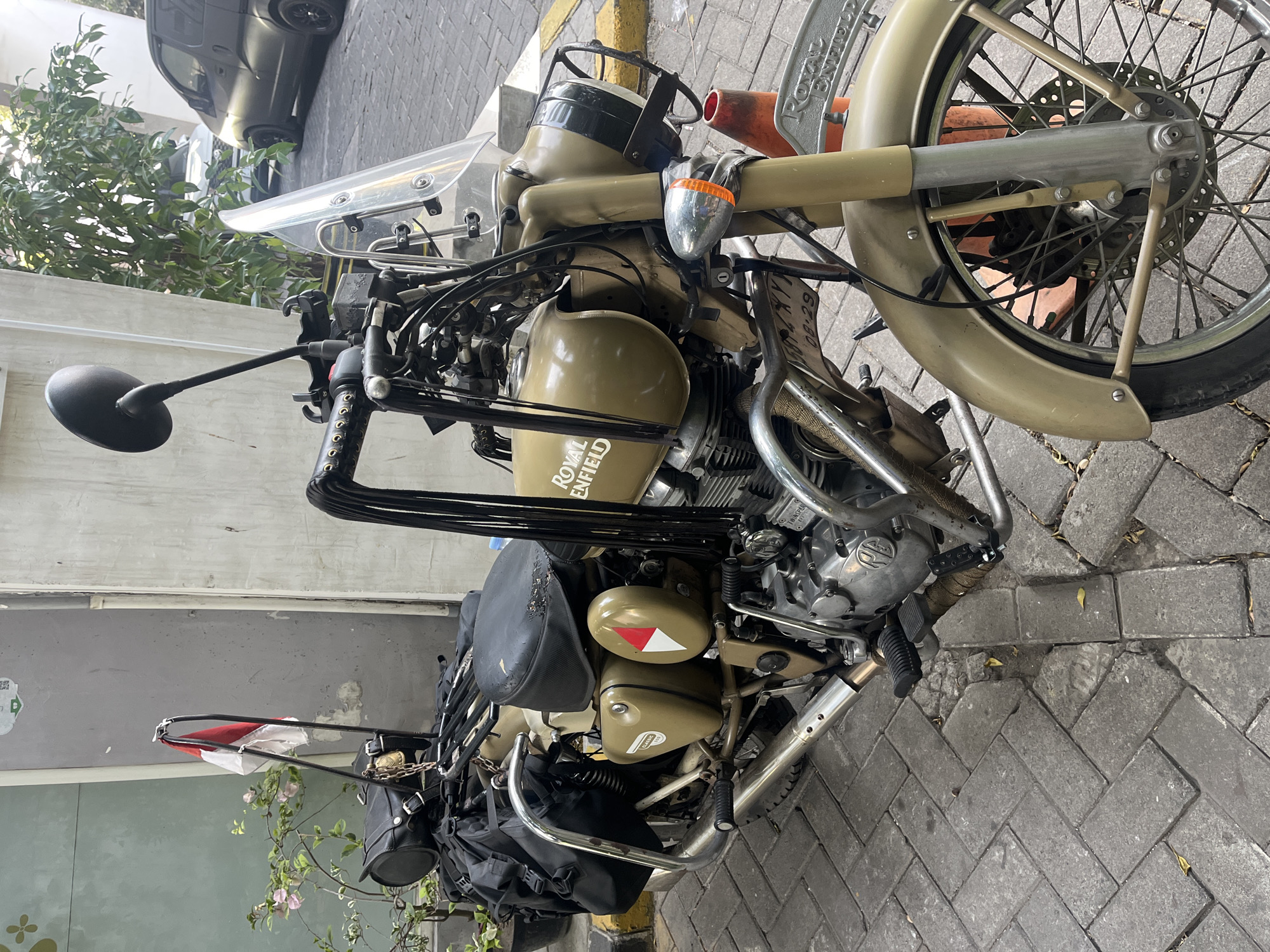 A tan Royal Enfield motorcycle is parked on a cobblestone street in Gapurosukolilo, Indonesia. The motorcycle is equipped with saddlebags and a windshield. A red and white Indonesian flag is attached to the back of the motorcycle. A dark-colored van is parked in the background, and a potted plant with pink flowers is visible to the left of the motorcycle. The scene is well-lit, suggesting it is daytime.