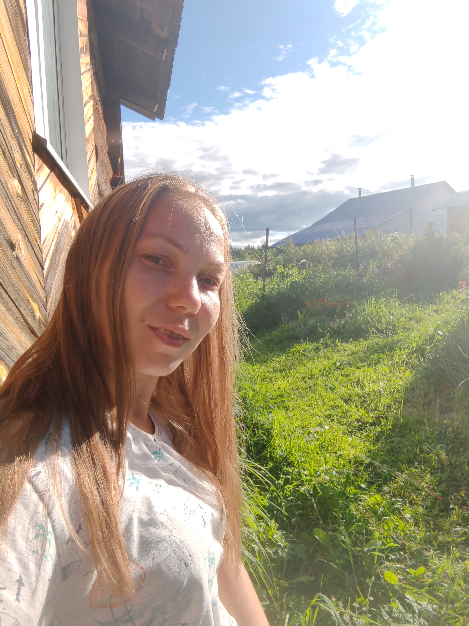 A young woman with long, reddish-blonde hair smiles at the camera, her face illuminated by the sun. She is wearing a light-colored t-shirt with a pattern of various nautical and tropical elements, including sailboats, palm trees, and birds. She is standing outdoors, with a wooden structure to her left, possibly a part of a house or shed. The background features a bright, sunny sky with scattered clouds, a green, grassy yard with some vegetation and flowers, and a building with a blue roof in the distance. The overall scene suggests a relaxed, outdoor setting, likely in a rural or semi-rural area. While specific location within Чухлэм, Russia is not evident from the image, the presence of a wooden house and abundant greenery is consistent with a Russian countryside environment.