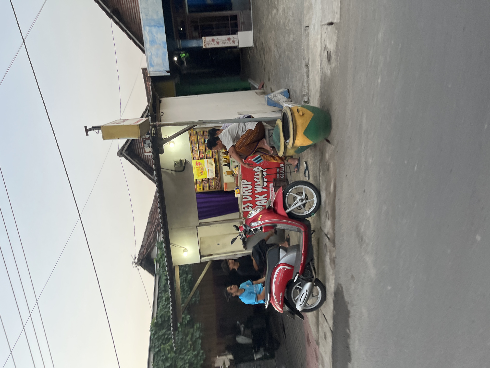 The image depicts a street scene in Karangtanjung, Indonesia, likely during the late afternoon or early evening due to the soft, diffused light. In the foreground, a red and silver scooter is parked on the side of a paved road. Two individuals are visible near the scooter. One, wearing a light blue shirt, is seated on the scooter, and another, wearing dark clothing, is standing behind him.
Behind the scooter is a small roadside stall or shop. A man, wearing a white shirt and a patterned sarong-like garment, is seated on a large, decorative object that resembles a stylized watermelon or gourd, painted green and yellow. He appears to be interacting with the stall. The stall itself has a canopy and displays various items, including what look like packaged goods and a prominent red banner with white text. Visible text on the banner includes "ES DROP," "JAK YAKIS," and "CARI ALL K." Another sign above the stall is yellow and appears to have text on it as well, but it is less legible. The building behind the stall has a corrugated metal roof and white walls, with some decorative elements. Green vines or foliage are visible growing on the side of the building and along a fence. Overhead, electrical wires crisscross the sky, which is a pale, hazy white, suggesting a cloudy or overcast sky. The overall impression is of a casual, everyday scene at a local vendor.