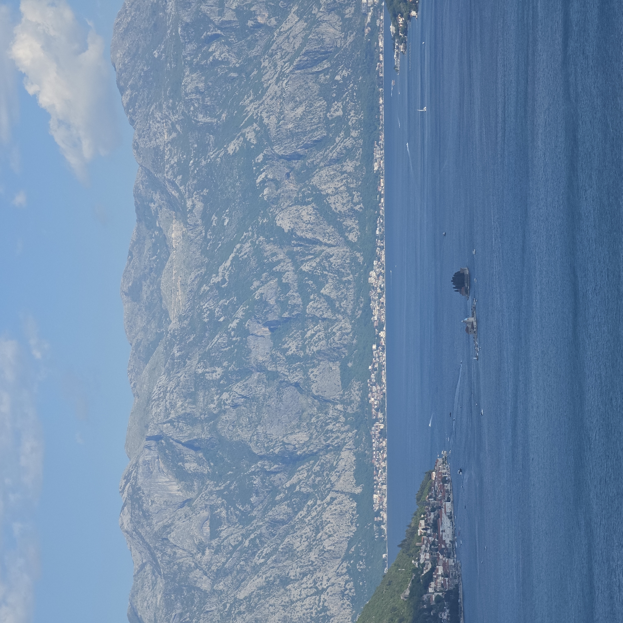 The image depicts the landscape around Morinj, Montenegro. A tall, rocky mountain range dominates the left side of the frame. The right side is occupied by a deep blue body of water, presumably part of the Bay of Kotor. A narrow strip of land runs between the mountain and the water, dotted with buildings that suggest a town or village. A small island with structures on it is visible in the water. Several boats dot the water's surface. The sky above the mountains is pale blue with scattered clouds.
