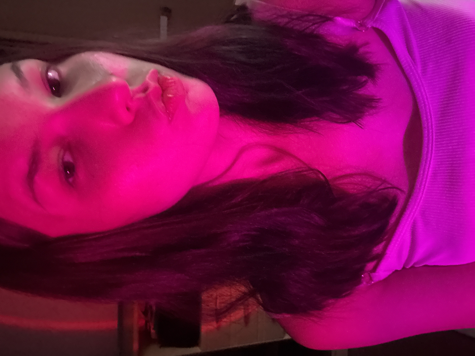 Inskoy, Russia. The image is a close-up, low-angle selfie of a young woman illuminated by pink light. She has dark hair and is wearing a white tank top. Her lips are pursed, and her gaze is directed at the camera. The background is dark and out of focus, with a hint of a wall on the right side.