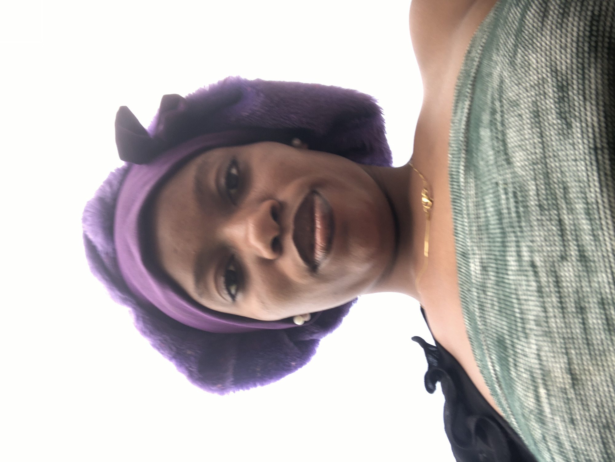 A low-angle, medium shot captures a person from the chest up, facing the camera. The person wears a purple headwrap with fluffy trim and small, dark adornments on either side, resembling ears. They also have a single pearl earring in their visible ear. The person's clothing consists of a black strap across the left shoulder and a green and white striped strapless top or dress. A thin gold necklace with a small pendant rests on their collarbone. The background is a bright, blown-out white, suggesting an outdoor setting with strong lighting, possibly during the day. The image is taken in Kashere, Nigeria.