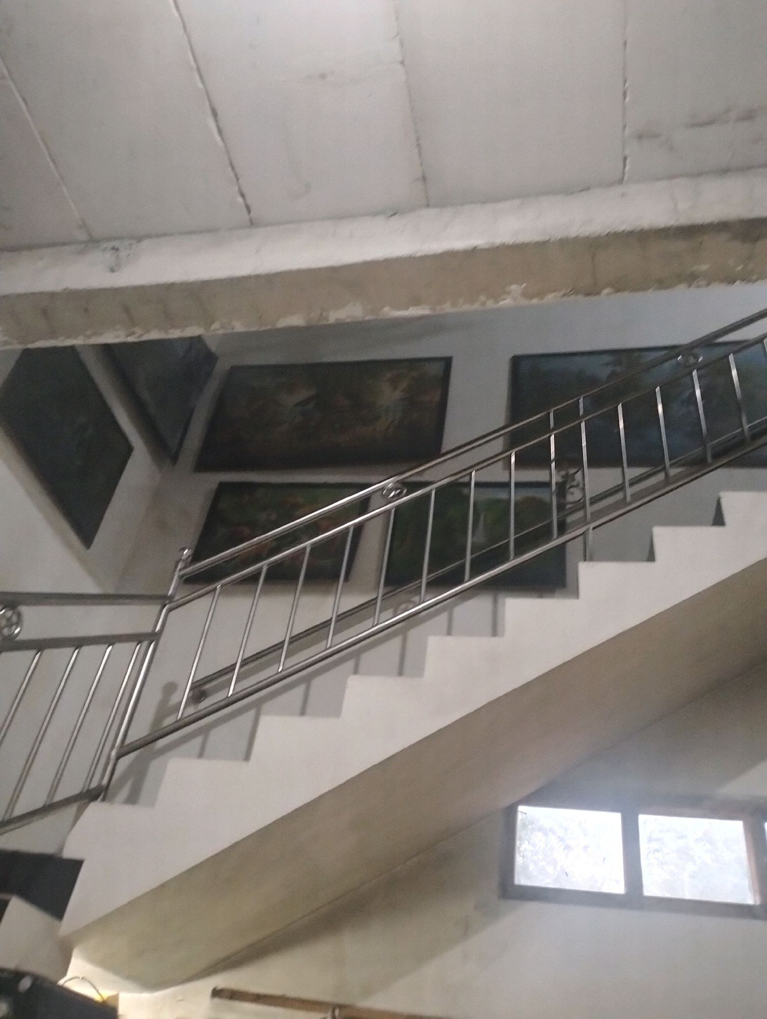 This image depicts an indoor staircase with a stainless steel railing. The staircase is made of concrete and painted white, with a light brown concrete beam running along its side. Several framed paintings are hung on the wall to the left of the stairs. Two small, rectangular windows with wooden frames are visible at the bottom right of the image. The ceiling appears to be made of concrete panels. The lighting suggests an indoor environment with some natural light coming from the windows. The overall scene is a functional interior space that also incorporates decorative elements. There are no people or animals visible in the image.