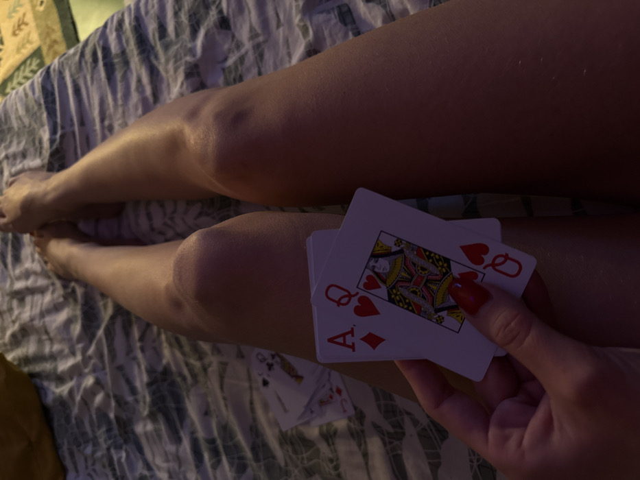 A person is sitting in bed with legs extended. They are holding a hand of playing cards, which include an Ace of Diamonds and a Queen of Hearts. Several additional cards are scattered on the bed. The bedding has a muted green and white patterned design. The lighting is dim, casting shadows across the legs and cards. A small section of mustard-colored fabric is visible at the edge of the frame. The focus is on the cards held in the hand.