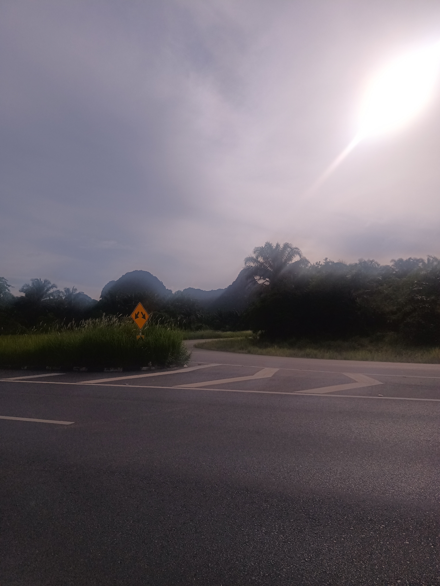 The image shows an asphalt road intersection on the outskirts of Ban Klang, Thailand, facing northeast. A yellow traffic sign with two arrows indicating directional choices is partially obscured by tall grass in the median. Behind the median, a landscape features a mix of palm trees and dense tropical vegetation, against the backdrop of a prominent, rounded mountain range shrouded in haze. The sky is overcast, with the sun positioned high and to the right, its light diffused by the clouds. The asphalt surface shows some wear and tear, with painted road markings delineating traffic lanes and directions. There are no people visible.