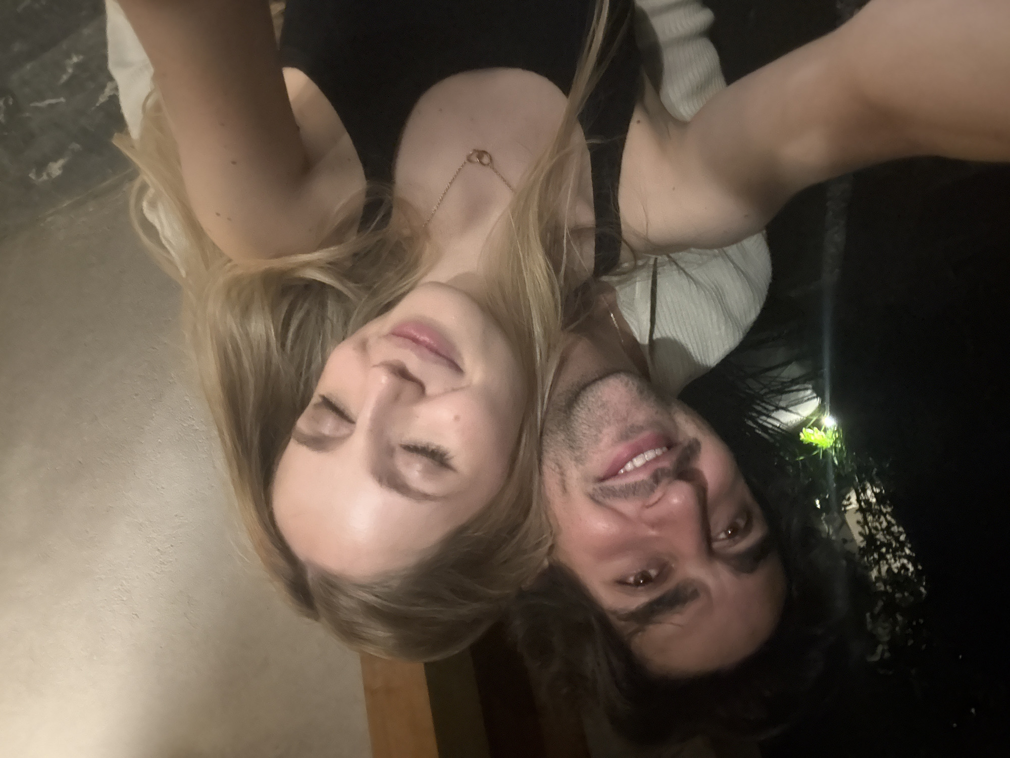 A selfie captures a fair-skinned woman with closed eyes and a blonde, shoulder-length hairstyle. She wears a black tank top and a delicate gold necklace. Beside her, a dark-haired, fair-skinned man smiles with an open mouth. He is wearing a white, textured sweater. The lighting suggests it is nighttime. Behind the man, there are dark shapes of trees. The location is Mexico City, Mexico.