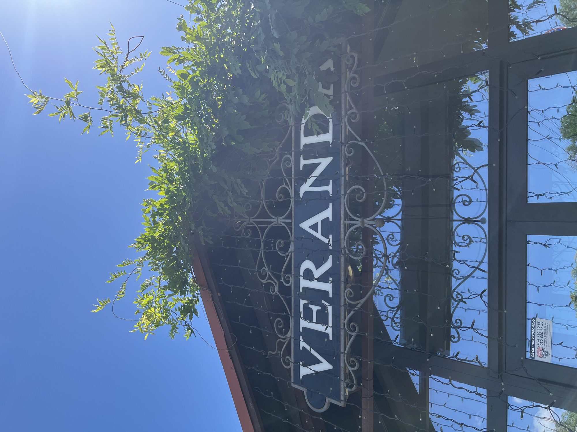 The image shows the entrance to a building with a sign that reads "VERANDA". The entrance is adorned with string lights and decorative metalwork. Green vines and leaves are growing over the top of the entrance. The sky is clear and blue, indicating a sunny day. There is a security sticker visible on the lower left window, which appears to be in Ukrainian and displays a phone number. The sticker has a crest with the text "ОБ'ЄКТ ПІД ОХОРОНОЮ" (Object under guard). The location is identified as Odesa, Ukraine.