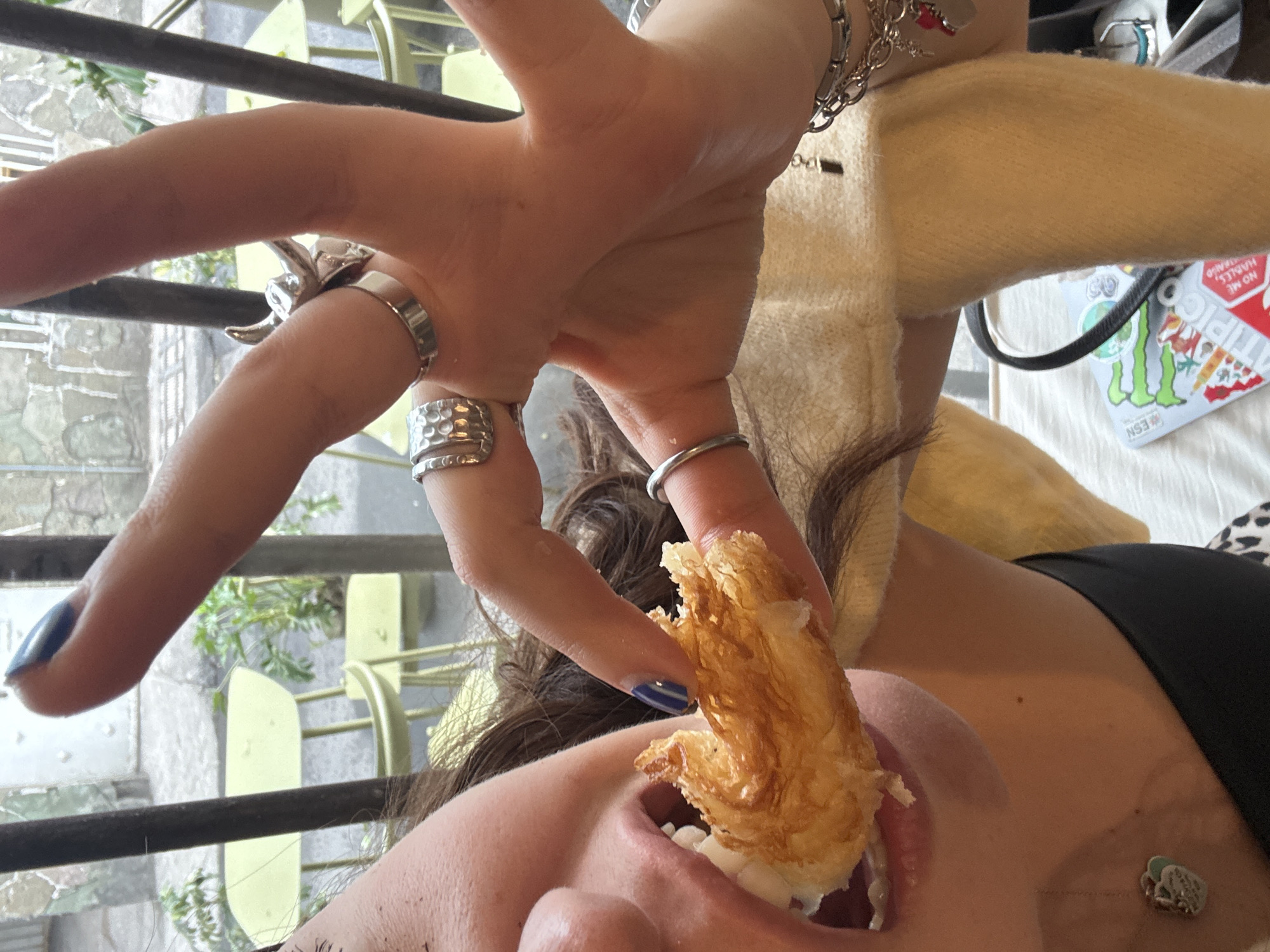 A person is holding a pastry to their mouth. The person has various rings on their fingers and painted nails. The location is possibly Mexico City, Mexico, based on an "epic" sticker.