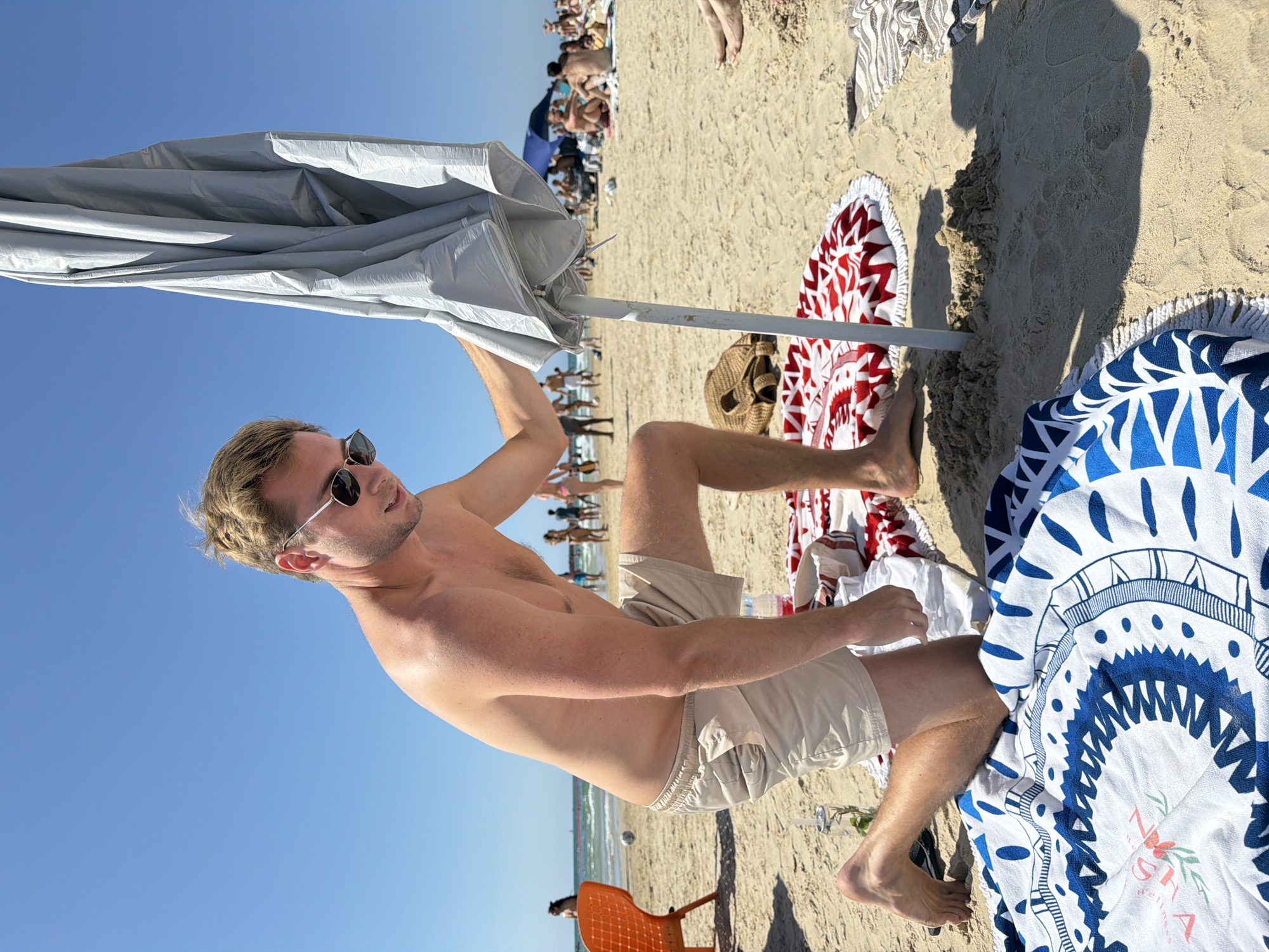 A light-skinned man with blond hair and sunglasses is depicted on a beach. He's shirtless, wearing tan shorts, and holding a large, open, silver-colored beach umbrella. The scene is sunny with a clear blue sky. Behind him, a sandy beach stretches out, populated with other beachgoers, some of whom are in the water. A red and white round beach towel is visible near his feet, along with a blue and white round beach towel. An orange plastic chair is located in the bottom of the frame. The location is Tel Aviv, Israel, identifiable by the beach environment and the presence of the Mediterranean Sea in the background.