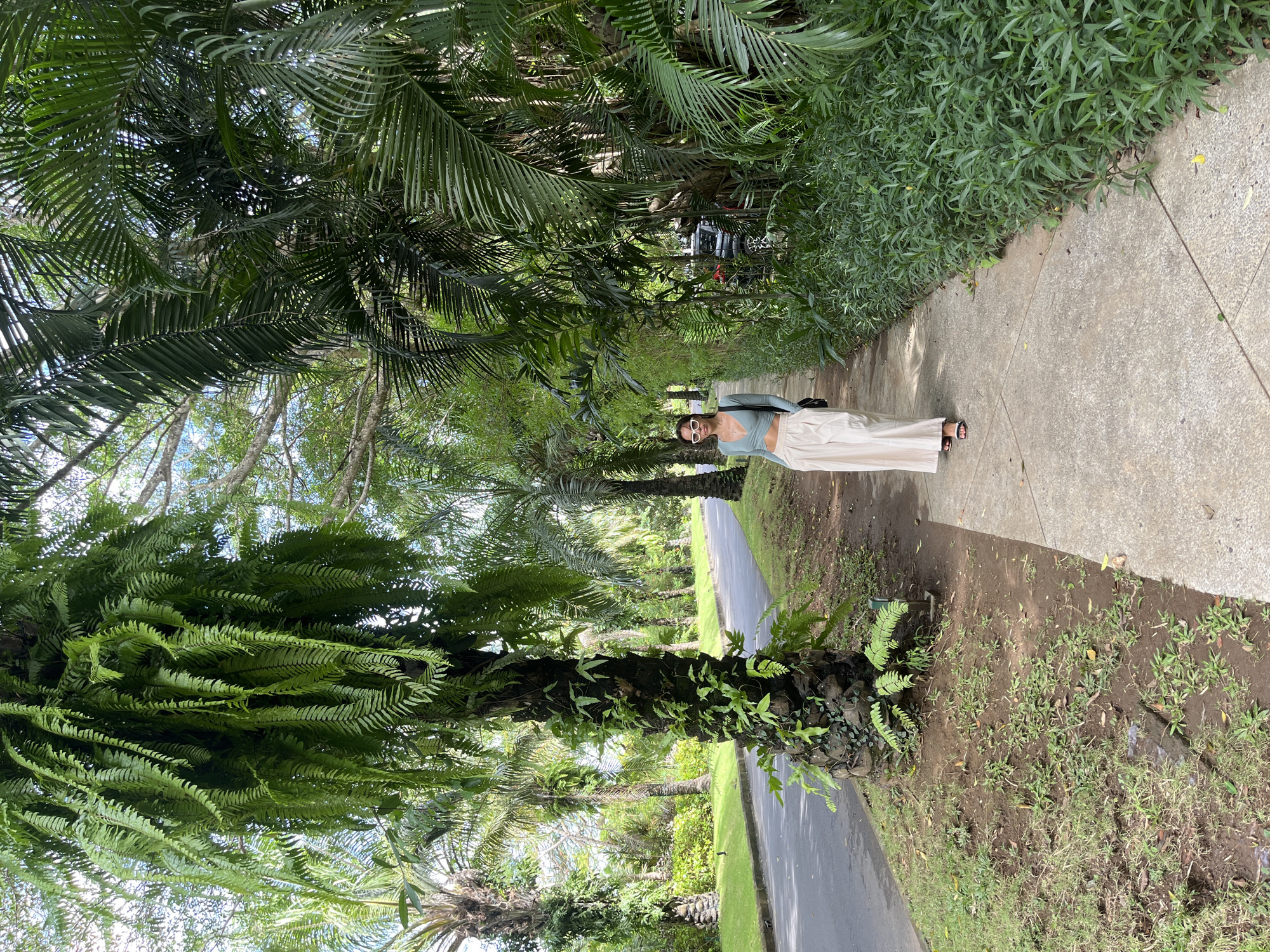 A full-length, outdoor photograph shows a woman standing on a sidewalk. She faces the camera, wears a light blue long-sleeve crop top, light beige wide-leg pants, and sunglasses. A black shoulder bag hangs from her right shoulder. She has bare feet in sandals. The scene is a sidewalk in a tropical location, possibly in Kuta Selatan, Indonesia. Lush, dense greenery, including palm trees and ferns, lines both sides of the sidewalk, creating a canopy overhead. A narrow road runs adjacent to the sidewalk. In the background, a vehicle is visible through the trees. The scene is illuminated by natural daylight.