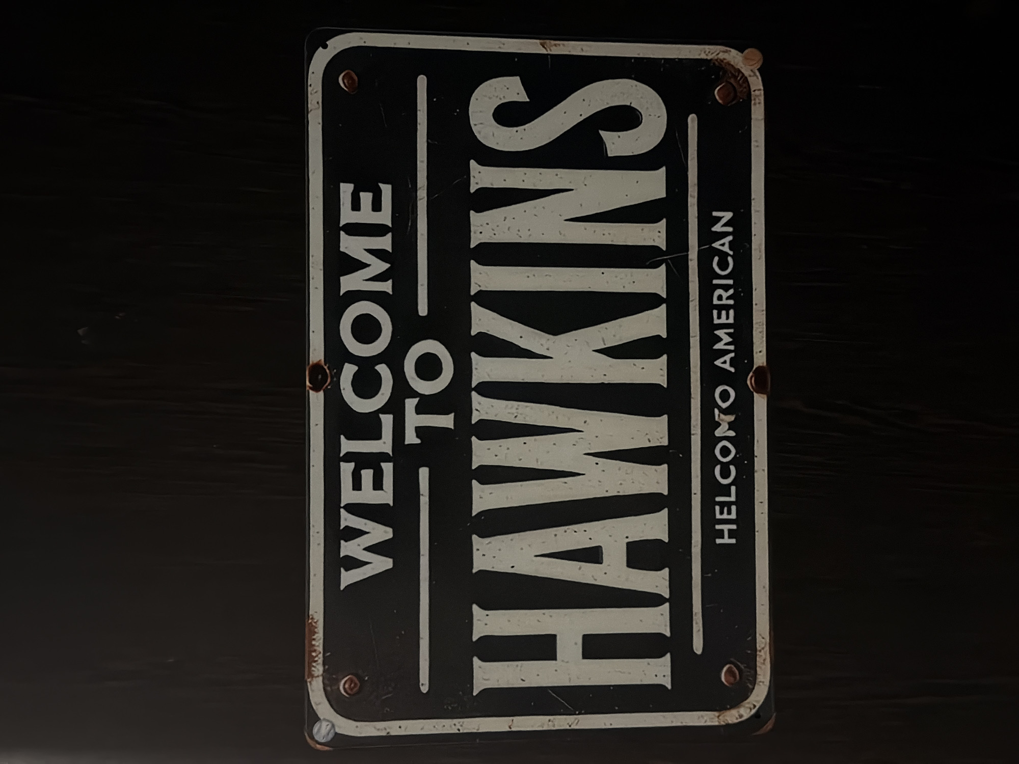 A rectangular, distressed metal sign with rounded corners, oriented vertically, rests against a dark, blurry background that appears to be a dark wooden surface. The sign is primarily black with white lettering and trim. The text reads "WELCOME TO HAWKINS" in large, bold, sans-serif font, stacked vertically. Below "HAWKINS", the sign reads "HELCOMO AMERICAN". Rust and wear are visible around the edges and on the lettering, giving the sign an aged appearance. There is no indication that the sign is located in Tbilisi, Georgia.