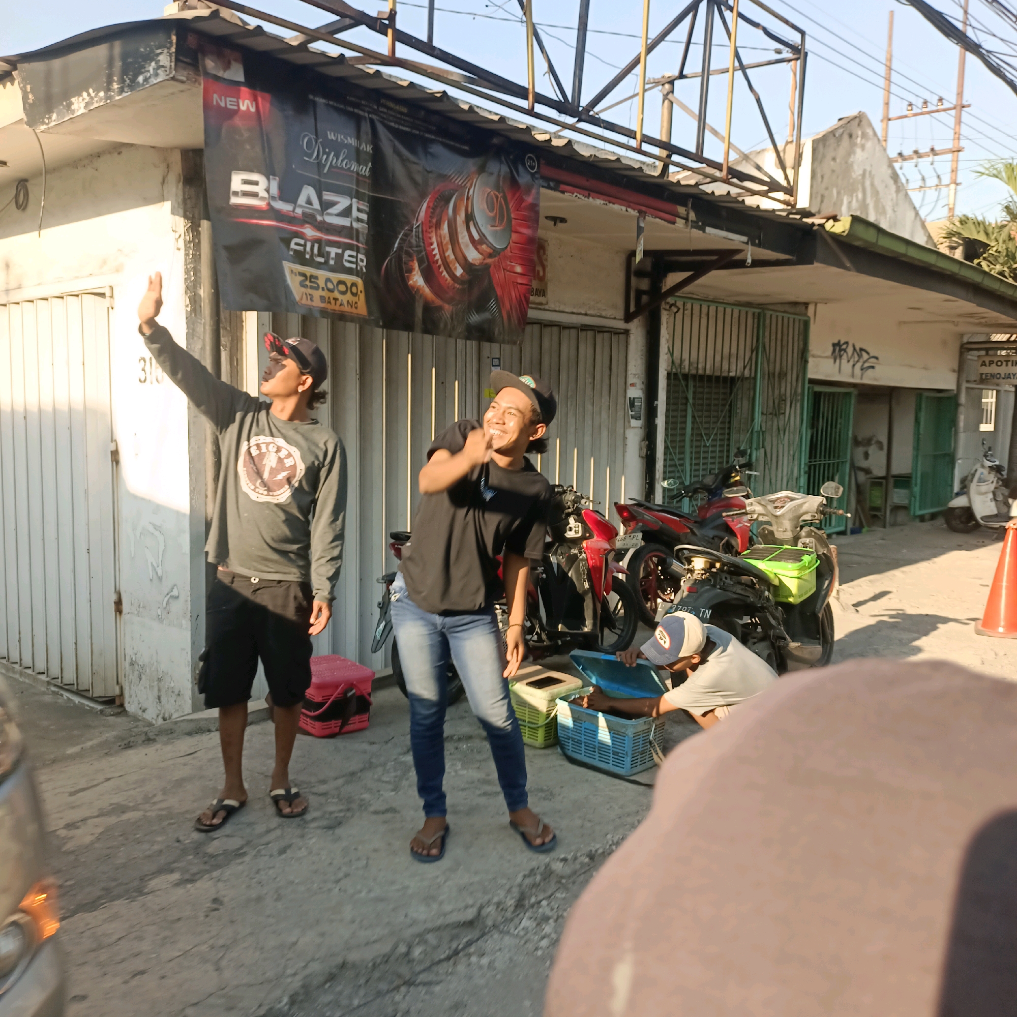 In Surabaya, Indonesia, on a sunny day, three young men are gathered outside a building with a corrugated metal roof. The building appears to be a shop or business, with a banner hanging above the entrance advertising "BLAZE FILTER." The banner features an image of a mechanical component.
The man on the left, wearing a grey long-sleeved shirt and black shorts, stands with his arm raised as if waving or signaling. He is wearing a black baseball cap and flip-flops. Next to him, a man in a black t-shirt and blue jeans is smiling and gesturing with his hands, also wearing a baseball cap and flip-flops. To the right, a third man, wearing a light-colored t-shirt and a baseball cap, is crouched down, looking into a blue plastic basket.
Several motorcycles are parked behind the men, including a red one and a silver scooter. In the foreground, a large, out-of-focus object, likely a shoulder or head, partially obstructs the view. The ground is paved with concrete, and there are some green baskets and red crates visible. In the background, parts of other buildings and utility poles are visible, suggesting an urban environment. The overall scene depicts a casual gathering outdoors, possibly during a break or while engaged in an activity related to the nearby motorcycles or the shop.