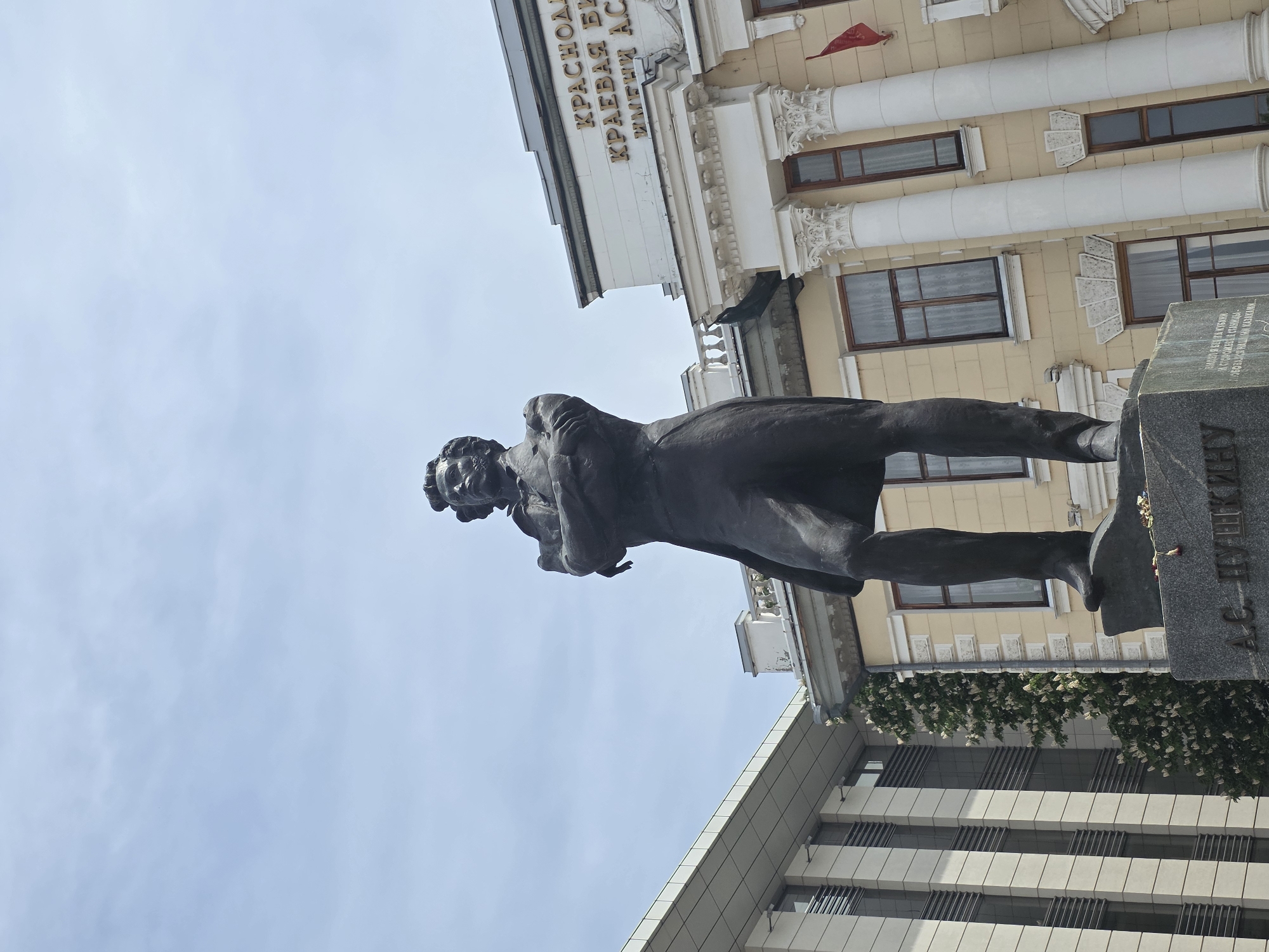 Here's a detailed description of the image:
The image is a low-angle shot of the monument to Alexander Pushkin located in Krasnodar, Russia. The bronze statue stands on a granite pedestal, with an inscription in Russian. The statue depicts Pushkin in a pensive pose, with a contemplative expression. The monument is situated in front of two buildings; an old, light-yellow building with white columns and windows, and a more modern grey building with gridded windows. The sky is visible in the background and is a pale blue color.