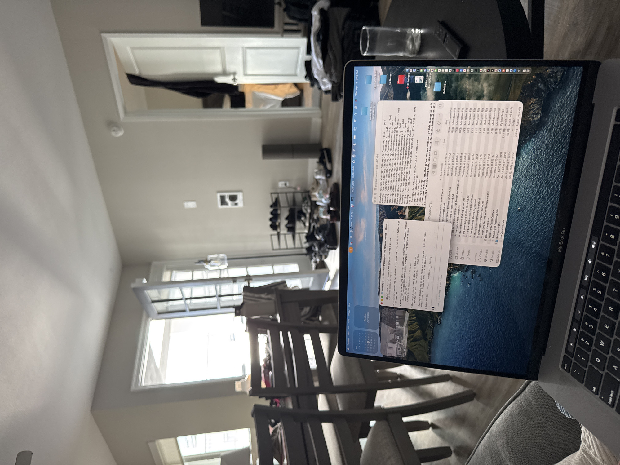 A low-angle, slightly canted indoor shot shows a MacBook Pro displaying a Finder window and a Terminal window, on a gray upholstered seat. The screen shows a list of markdown files in Finder and a command-line interface in Terminal. To the left, through a doorway, is a well-lit interior space with a window that lets in daylight. The house appears modern, with simple paint colors and trim work. In the background, there are indications of home life: clothing, shoes, a table. The scene evokes a sense of working from home in a comfortable, modern residential setting. I cannot confirm the location in the city of Redondo Beach, United States of America.