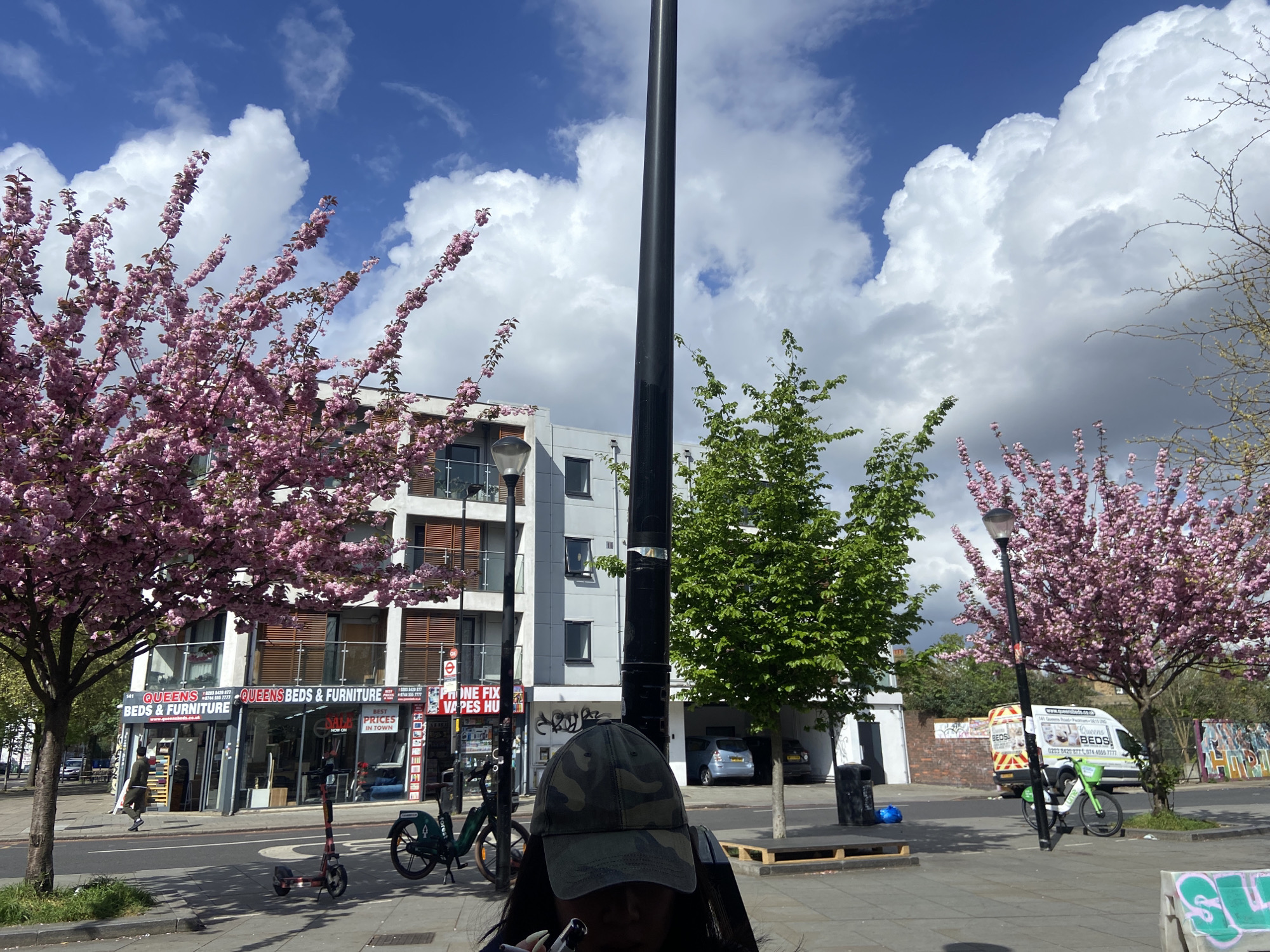 Here is a detailed description of the image:
**Scene:** The image shows a street scene in London, United Kingdom, on a day with blue skies and scattered clouds. Blossoming trees with pink flowers frame the view on both sides.
**Buildings and Businesses:** On the left is a commercial building housing "Queens Beds & Furniture" and "Phone Fix/Vapes Hub" visible from the street. The building has multiple floors with apartments above the storefronts.
**Street Furniture and Transportation:** The foreground shows a paved area, possibly a sidewalk. There are parked Lime bikes and electric scooters. A light pole is situated in the center of the frame. A white van is parked on the right side of the street.
**People:** A person wearing a camouflage cap partially obscures the view in the foreground. Another person is walking into the "Queens Beds and Furniture" store.
**Environment:** A patch of green foliage and trees is noticeable. A wall with some graffiti is also visible. The sky is predominantly blue with prominent white clouds.