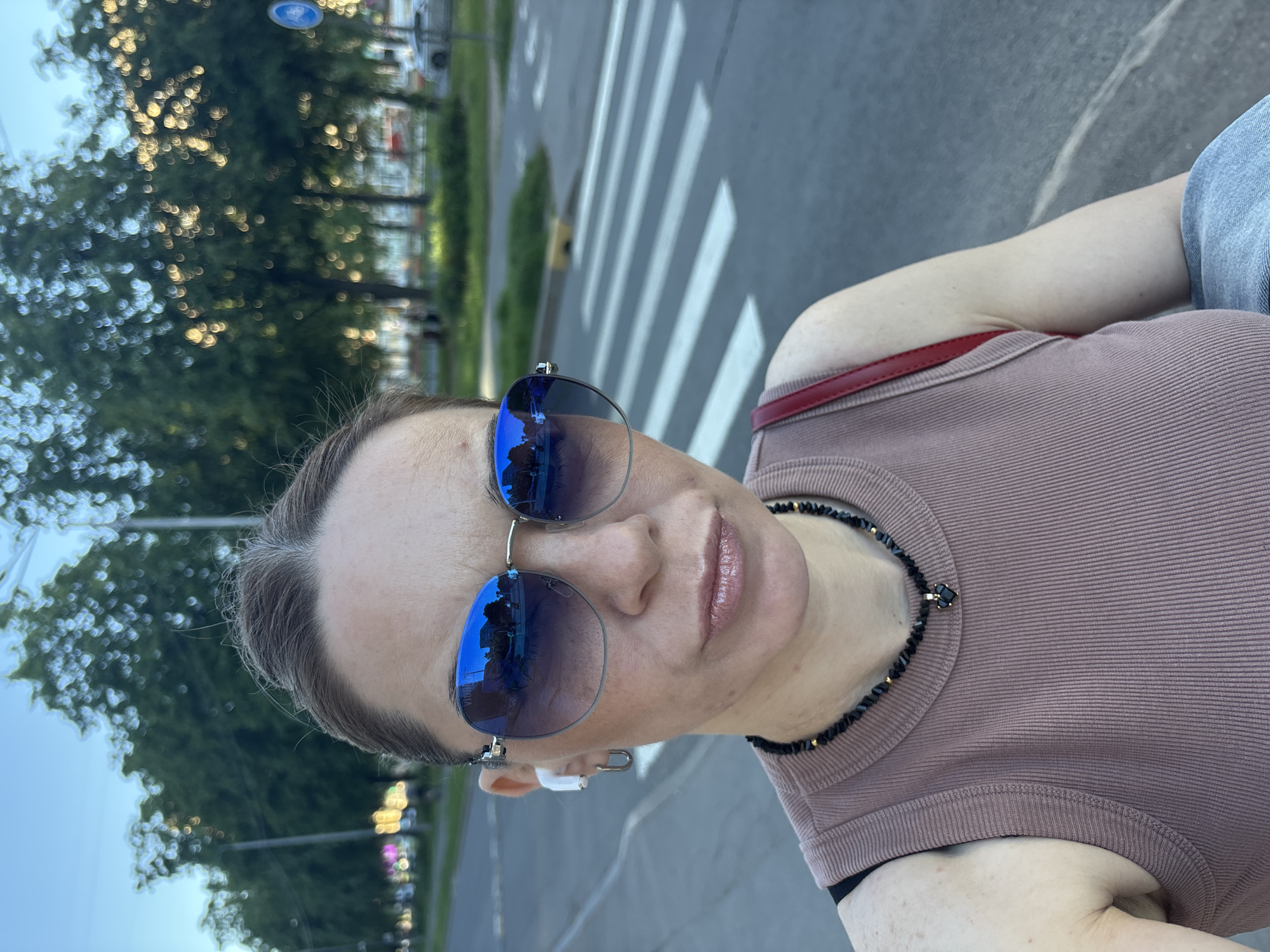 A middle-aged woman is captured in a close-up, selfie-style outdoor photograph in Sinyavino, Russia. She faces the camera directly, her eyes concealed by large, blue mirrored sunglasses with silver-toned frames. The left lens of the sunglasses features the visible brand name "VIBRO." A white wireless earbud is inserted in her left ear, complemented by a silver-toned rectangular hoop earring. Her hair is pulled back, revealing visible grey strands on top. Her lips are slightly pursed and appear to have a light gloss.
She wears a ribbed, light brown or mauve tank top, with a black strap (likely from an undergarment) visible on her left shoulder. Around her neck, she has a black beaded necklace with small gold-toned accents and a central black heart-shaped pendant. A red strap, presumably from a bag, is visible over her right shoulder. A light blue denim garment is draped over her left arm.
The background depicts a blurred urban street scene. A prominent pedestrian crossing with white "zebra" stripes is visible on the dark grey asphalt road to the right of the woman. A blue circular road sign with a white bicycle symbol, indicating a bicycle path or lane, is situated further down the road on the right. A line of dense green trees, exhibiting strong bokeh effects from filtering sunlight, runs along the left side of the background. Faint, blurred lights are visible in the distance on the left, possibly from streetlights or buildings. On the far right, a light-colored vehicle and indistinct building structures are visible. The overall lighting suggests natural daylight, likely late afternoon or early evening.