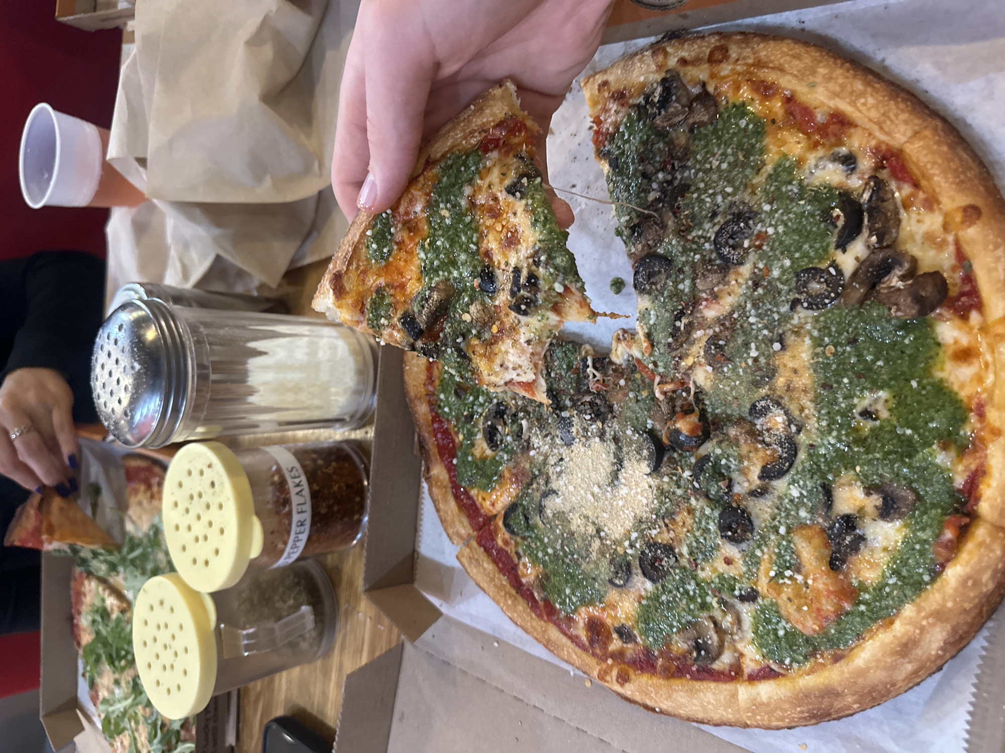 This image depicts a casual indoor dining scene, likely in a pizza restaurant or casual eatery in College Park, United States of America. The central focus is a round pizza, from which a hand is lifting a slice. The pizza is generously topped with a vibrant green pesto sauce, sliced black olives, and mushrooms, with a base layer of red tomato sauce visible beneath. A fine dusting of grated cheese and red pepper flakes is sprinkled across the pizza. The crust is thick, golden, and appears well-baked.
The hand lifting the pizza slice shows visible nail polish and a ring. Strands of melted cheese stretch between the lifted slice and the rest of the pizza, indicating it is freshly served and hot. On the wooden table, several condiment shakers are visible: one filled with grated white cheese (likely Parmesan), another with "PEPPER FLAKES" clearly labeled on its side, and a third containing dried green herbs. A white disposable cup and a brown paper bag are also present in the upper left corner of the frame. In the lower-left background, another person's hand and a section of a second pizza, topped with green leafy vegetables (possibly arugula), are visible, suggesting a shared meal among companions. The lighting is soft and uniform, typical of an indoor setting.