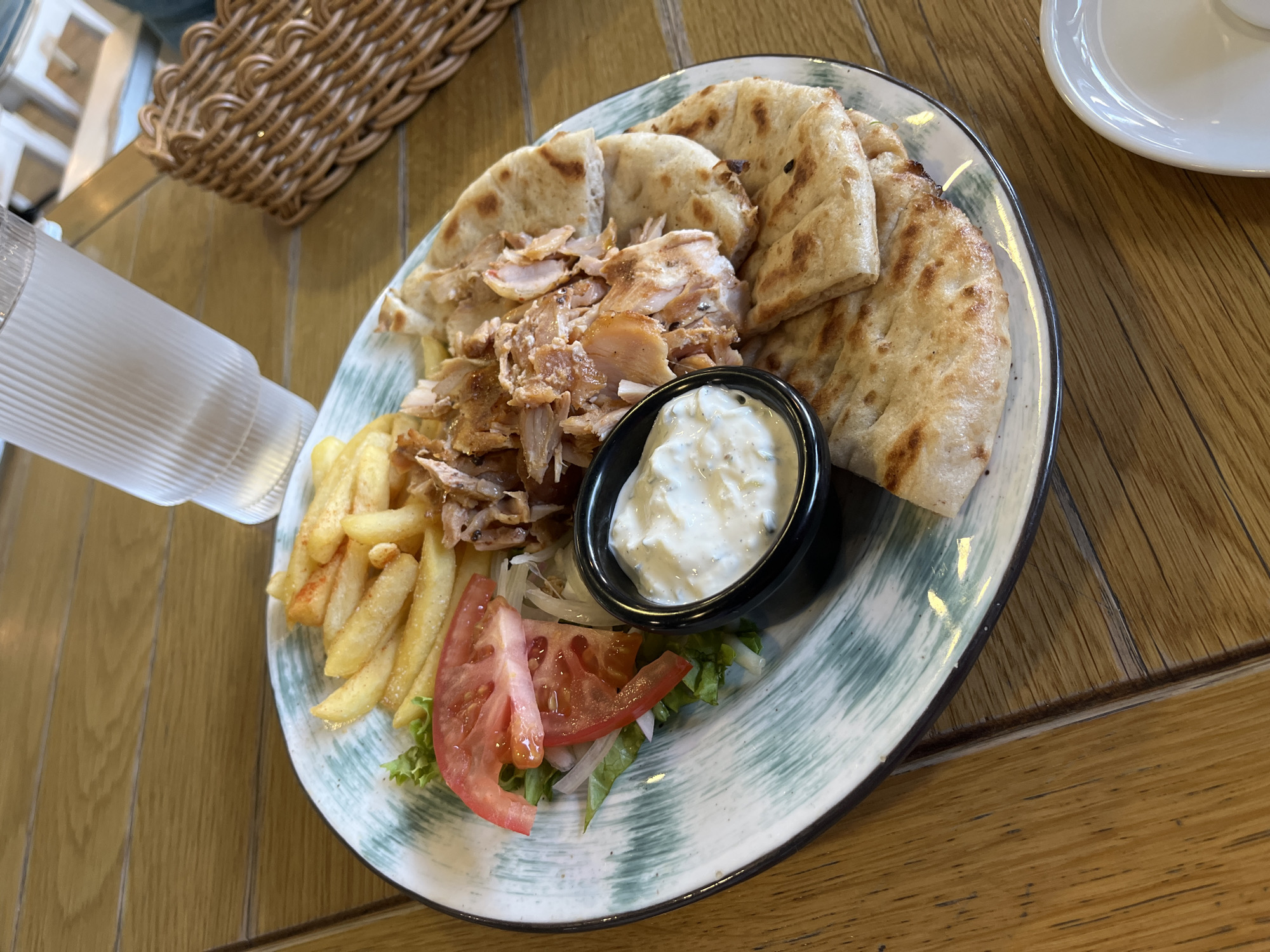 The image displays a meal served on a circular ceramic plate, situated on a light brown wooden plank table within an indoor environment in Sochi, Russia. The plate, featuring a light background with a teal and white abstract brushstroke pattern along its rim, contains several food items. Shredded, cooked meat, consistent with gyros, occupies the central-left area. A portion of golden-brown french fries is positioned at the top-left. Multiple pieces of grilled flatbread are arranged on the right side of the plate. A small side salad, comprising green leaf lettuce, two slices of red tomato, and thinly sliced white onion, is located on the left side of the plate. A small, black cylindrical ramekin containing a white, creamy sauce is placed between the salad and the shredded meat. In the upper right section of the frame, a clear, vertically ribbed glass filled with water is present. A light brown woven wicker basket is partially visible behind the glass and to its right. At the bottom-right corner of the image, a small, empty white ceramic dish is discernible. No individuals are depicted in the scene.