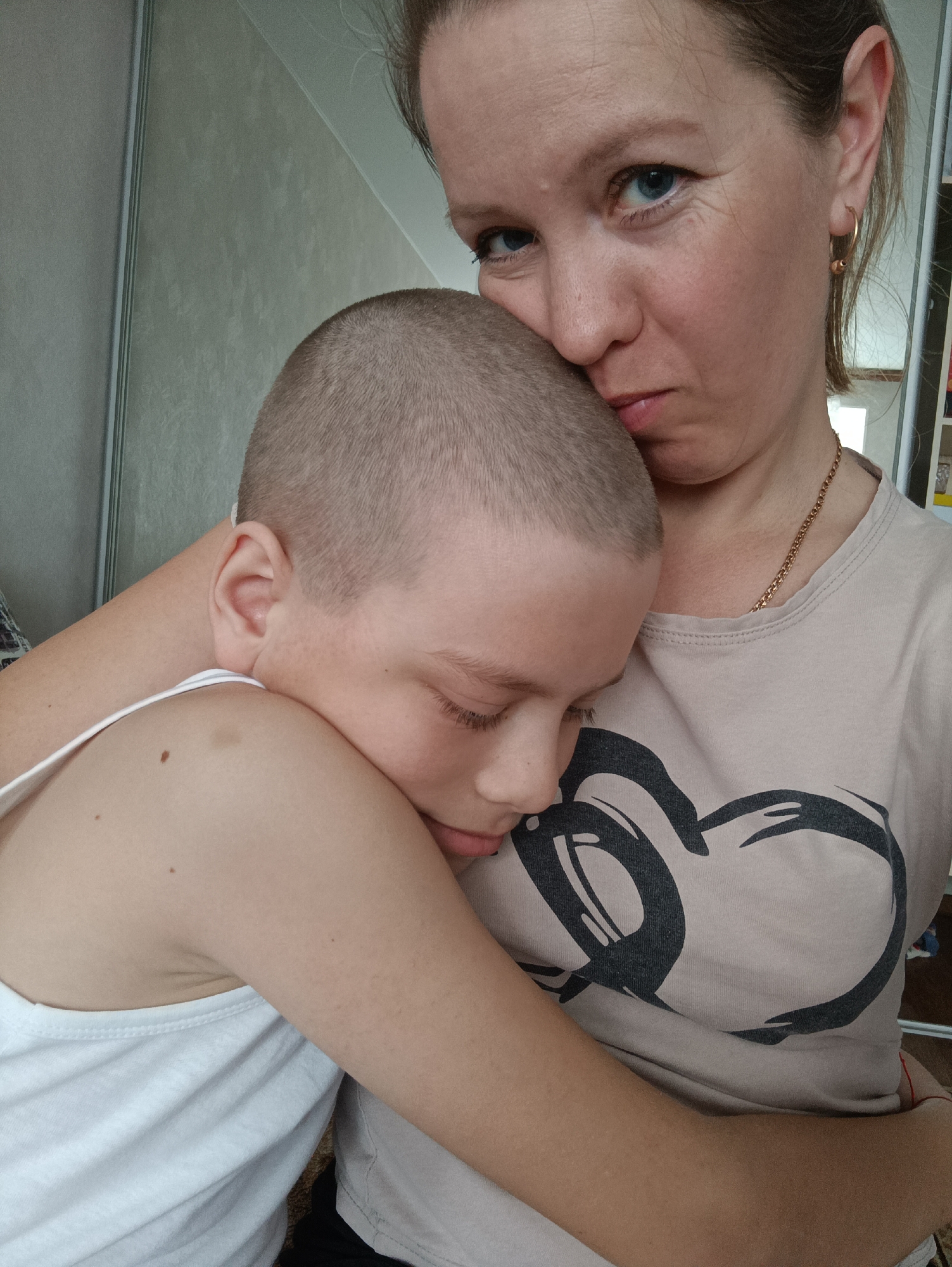 A medium shot captures a woman embracing a child with a shaved head. The woman, fair-skinned with blue eyes, is wearing a beige t-shirt with a black heart-shaped design and a gold necklace. She is looking directly at the camera with a slightly pensive expression. The child, fair-skinned with closed eyes, is wearing a white tank top and leaning into the woman's embrace. The location is likely inside a home, evidenced by a gray wall and partial view of a cabinet in the background. Given the user's prompt, the location is potentially in the city of Chaikovsky, Russia.