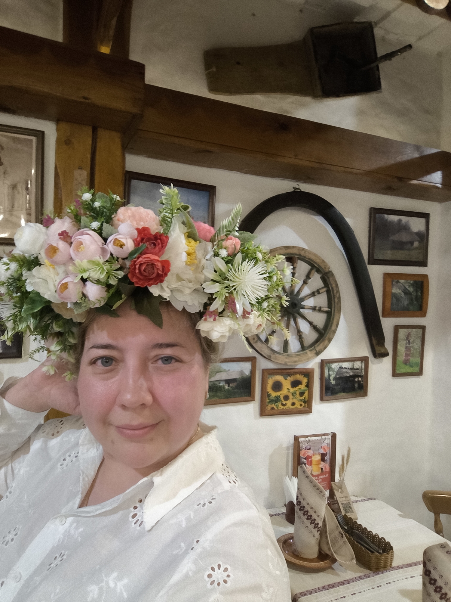 A woman wearing a large floral headpiece is centered in the image, slightly to the left. She is fair-skinned with blue eyes, and is wearing a white, eyelet button-down shirt. Her right hand is raised near her head. The background is the interior of a room, possibly a restaurant. Behind the woman are a variety of framed pictures and decorative items hanging on a white wall. These include landscapes, a picture of sunflowers, an old wagon wheel, and a dark, curved wooden piece (possibly part of a plow or carriage). A wooden beam is visible along the top of the wall. In the lower right corner, a table is partially visible, set with a white tablecloth, napkins, utensils in a basket, and a menu. Given the style of the headpiece, traditional clothing, and the interior decor, it is likely the image was taken in a restaurant or cultural center within Moscow, Russia showcasing traditional Russian or Eastern European culture. There is no evidence to determine a specific location in the city.