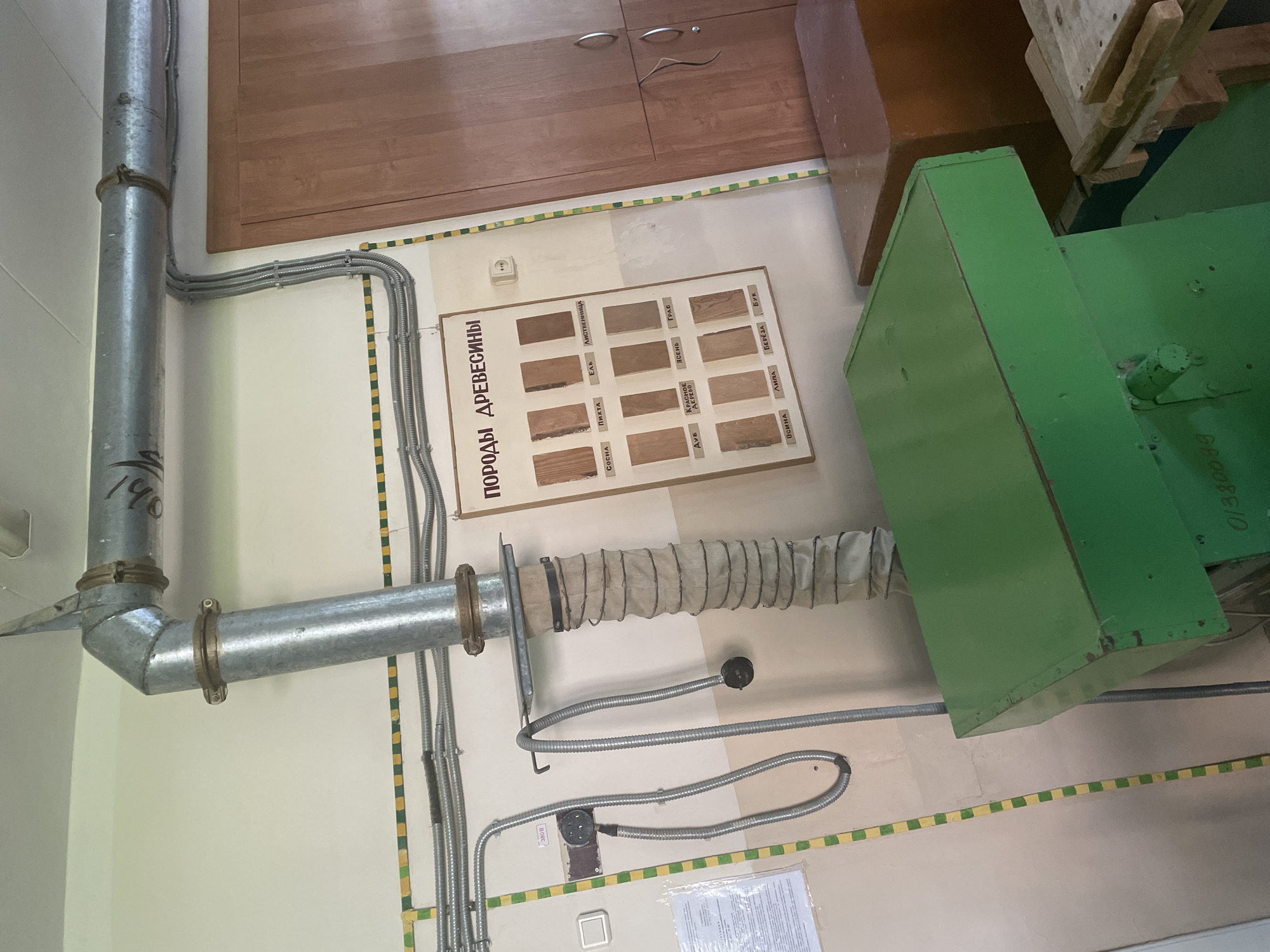 The image depicts an interior scene, likely a workshop or classroom, located in Chaikovsky, Russia. The primary focus is a large, green industrial machine positioned in the foreground, with a visible serial number or code "0/330099" inscribed on its side. Above and behind the machine, a large metal pipe system, presumably for ventilation or dust collection, descends from the ceiling. Adjacent to this pipe system is a bulletin board displaying samples of different wood types, identified by Russian labels such as "СОСНА" (Pine), "ПИХТА" (Fir), "ЕЛЬ" (Spruce), "ЛИСТВЕННИЦА" (Larch), "ДУБ" (Oak), "КРАСНОЕ ДЕРЕВО" (Mahogany), "ЯСЕНЬ" (Ash), "ГРАБ" (Hornbeam), "ОСИНА" (Aspen), "ЛИПА" (Linden), "БЕРЕЗА" (Birch), and "БУК" (Beech). The bulletin board is mounted on a light-colored wall, adorned with horizontal yellow and green striped tape along the upper and right edges. To the right of the bulletin board and extending to the edge of the frame is a large, light-brown wooden cabinet with two doors, each featuring two oval metal handles. Electrical conduits and wiring are visible throughout the scene, running along the walls and near the pipe system. A white electrical outlet with three circular holes is mounted on the wall. The environment appears to be functional and utilitarian, with evidence of industrial or educational equipment. There are no people visible in the image.