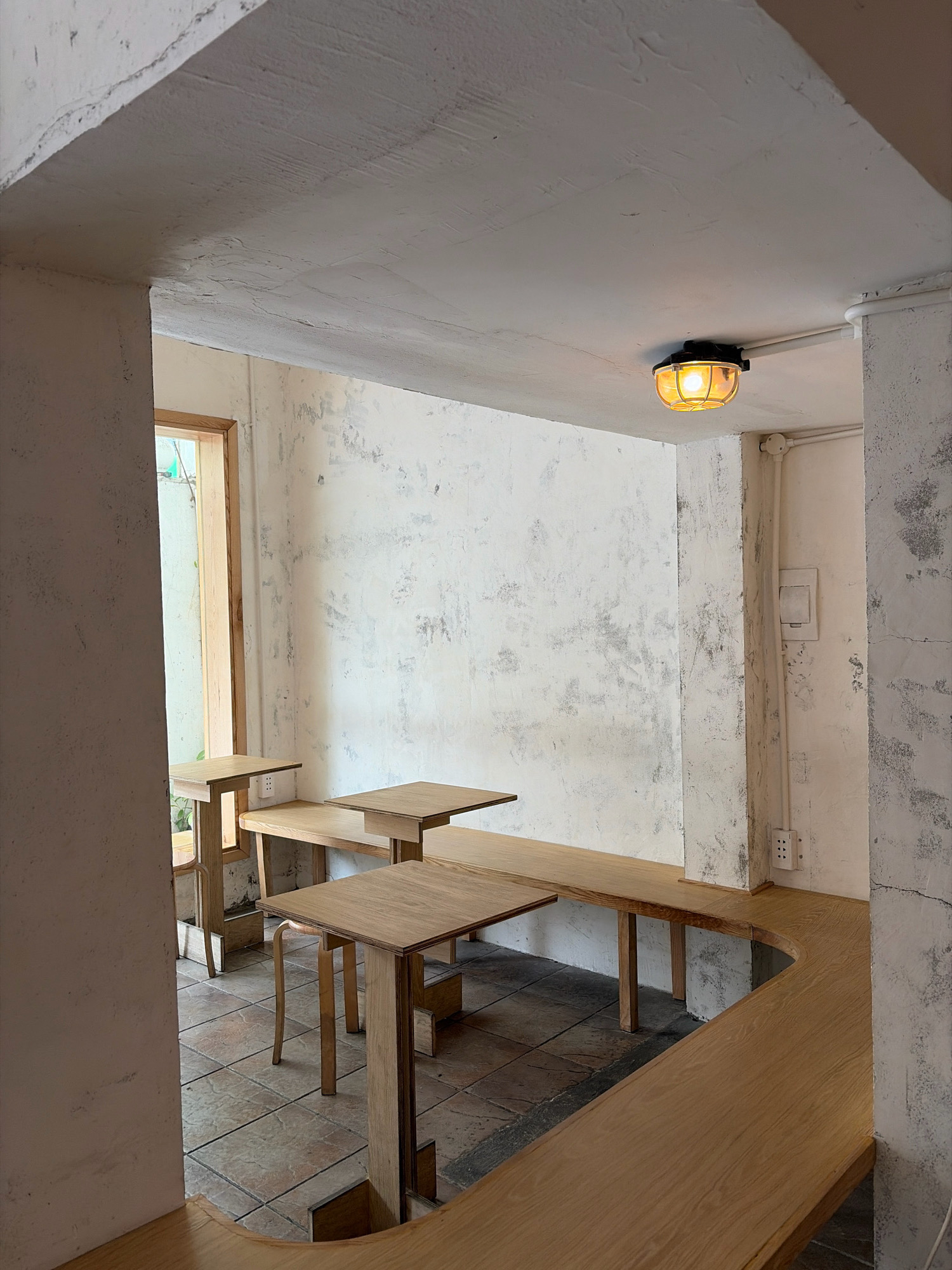The image depicts the interior of a cafe in Đà Nẵng, Vietnam. The cafe features a minimalist design with raw concrete walls and ceiling. Wooden tables and benches are strategically placed throughout the space, creating a cozy and inviting atmosphere. The lighting is provided by a single industrial-style pendant lamp with a yellow glow. A large window on the left side of the image offers a glimpse of the outside, suggesting an urban setting. The floor is tiled with a rustic pattern, adding to the cafe's unique aesthetic. The overall ambiance is calm and contemplative, ideal for enjoying a cup of coffee or tea.