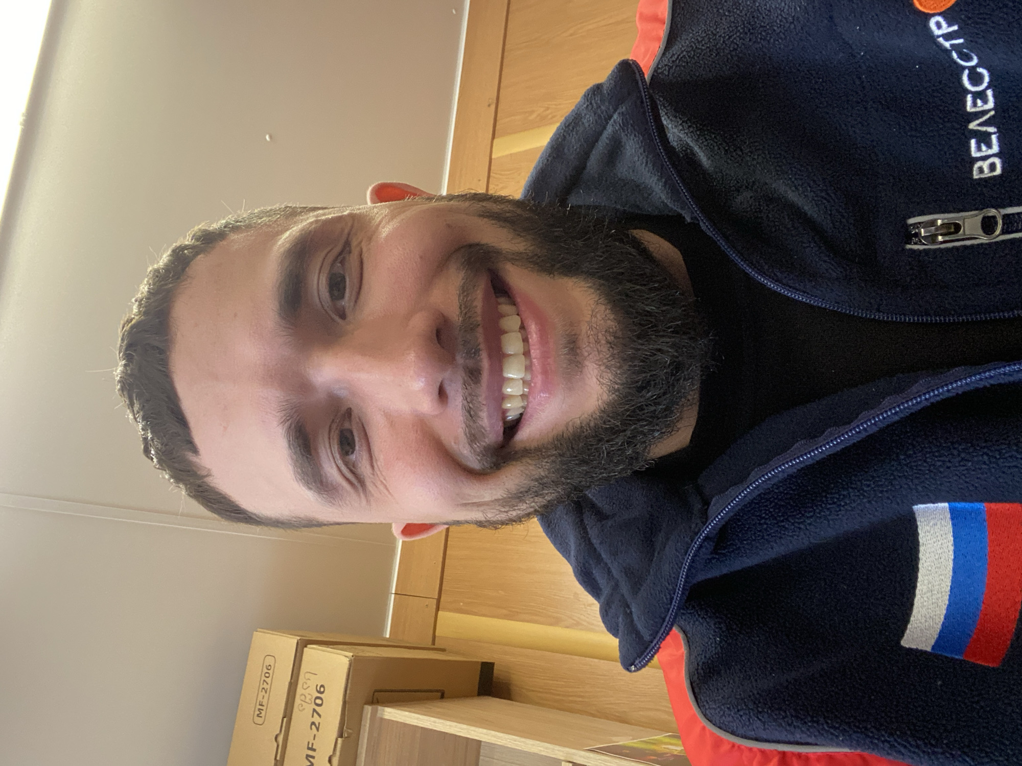 The image captures a smiling man in a close-up, selfie-style portrait. He has dark hair and a full beard, looking directly at the camera with a broad, toothy grin. He is wearing a dark blue fleece jacket with red accents. On the right side of his jacket (viewer's left), orange Cyrillic text reading "ВЕЛЕССТРОЙ" (VELESSTROY) is visible. On his left arm or shoulder (viewer's right), there is an embroidered patch depicting the Russian flag with its horizontal white, blue, and red stripes.
The background suggests an indoor setting, with light-colored walls or ceiling and light wooden furniture, possibly shelves or a desk. In the lower left corner of the frame, several cardboard boxes are stacked, one of which clearly displays "MF-2706" as part of its printed label. The man's cheerful expression and the presence of the Russian flag on his attire are notable details, aligning with the given location context of Luzhitsy, Russia.