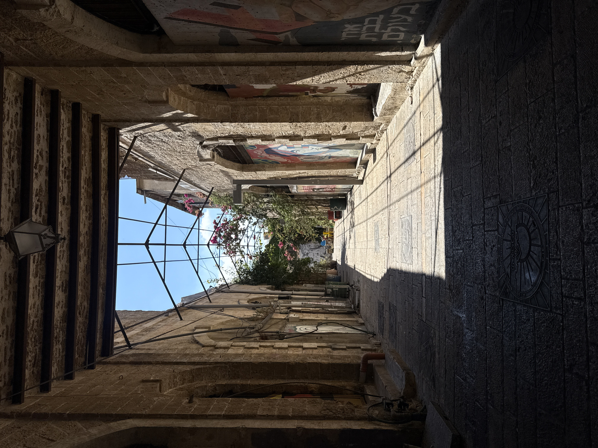 The image depicts a sun-dappled alleyway in Tel Aviv, Israel, likely within the ancient port city. The narrow passage is flanked by weathered stone buildings with arched entrances, some of which feature colorful murals. A metal frame structure, partially covered with vines and pink bougainvillea, extends overhead, casting a grid of shadows on the cobblestone pavement. The ground is paved with large, irregular stone blocks, and at least two circular metal grates with intricate designs are visible. The alleyway recedes into the distance, where more greenery and a hint of blue sky can be seen. There are no people visible in the frame. The lighting suggests it is daytime, with strong sunlight creating high contrast between illuminated and shadowed areas. The overall impression is one of historical charm and artistic expression within an urban setting.