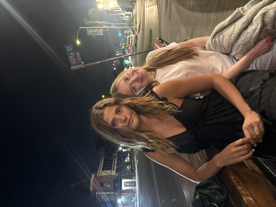 The scene depicts two young adult females seated outdoors at night, situated at the intersection of Vista Street and Larchmont Boulevard in Los Angeles, California, United States. This location is confirmed by a visible street sign displaying "Vista St" and the distinctive Tudor-style architecture of a building in the background.
The female on the left has long, wavy light brown hair and wears a black V-neck dress; she looks directly into the camera with a neutral expression, her left hand resting on a wooden surface beside a dark handbag. The female on the right has long, straight light brown hair and wears a white short-sleeved t-shirt; she smiles at the camera, leaning her head against the left female's shoulder, and holds a grey knitted garment on her lap.
In the background, to the left, stands a two-story building featuring Tudor-style half-timbering, a brick base, and a prominent conical-roofed turret. To the right, a commercial street stretches into the distance, illuminated by streetlights and vehicle headlights/taillights, with palm trees and additional commercial structures lining the thoroughfare. The sky is dark, indicating a nighttime setting.