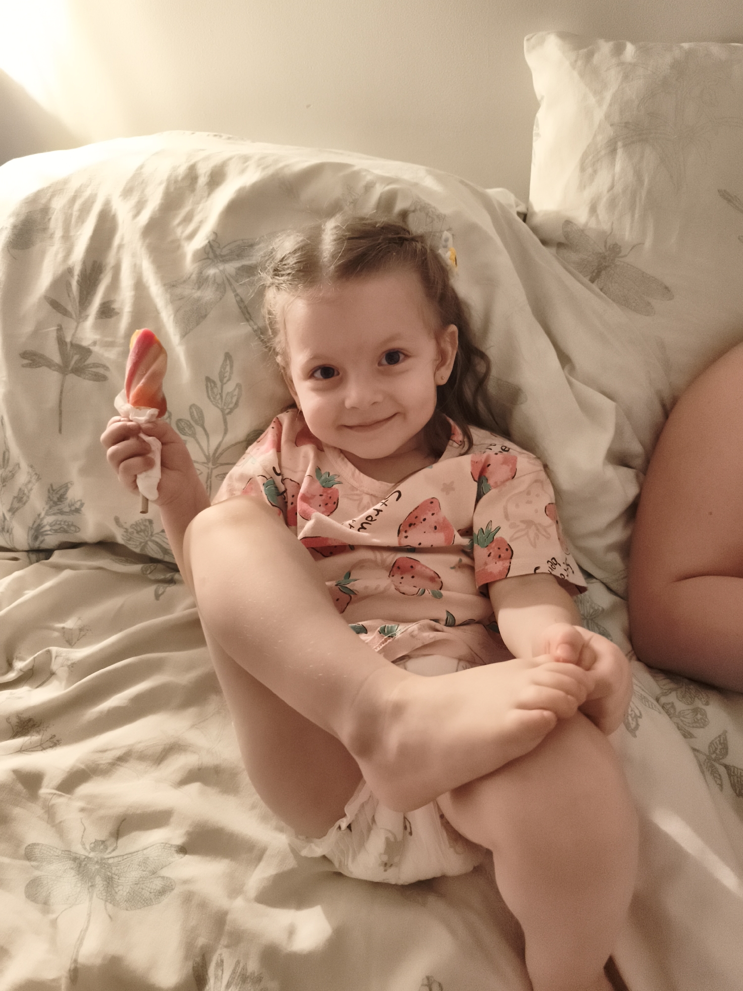 A young girl with brown hair tied in pigtails is lying on a bed covered with a patterned duvet. She is wearing a pink t-shirt with a strawberry pattern and a diaper. She is holding a multi-colored popsicle in her right hand and smiling at the camera. Her legs are crossed, with her right leg on top of her left. The background is a plain wall with some lighting creating shadows. The overall mood is relaxed and happy. The visible text on the t-shirt reads "merci". The scene appears to be indoors, possibly in a bedroom, and the time of day is likely evening or night due to the lighting.
